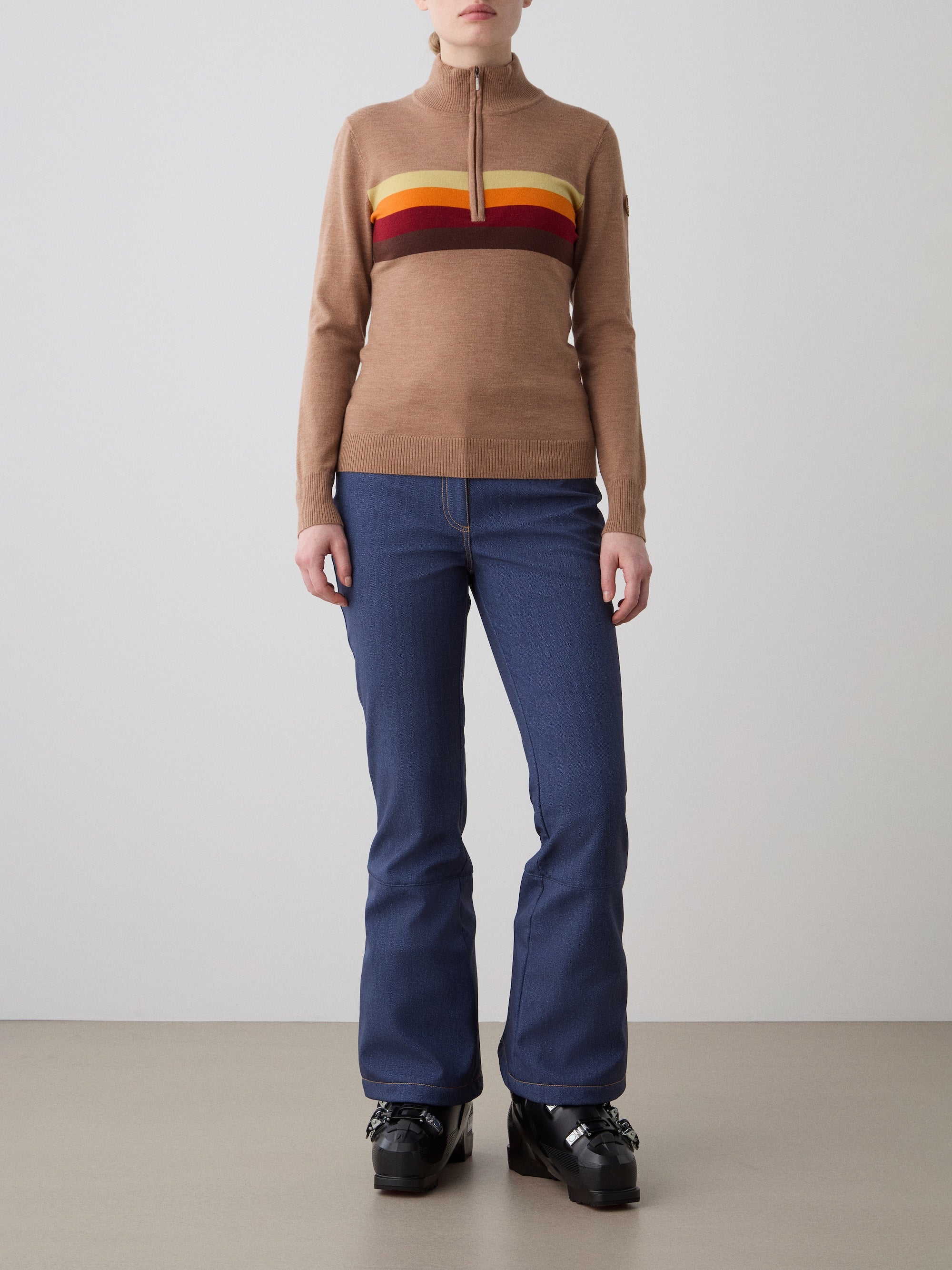 A woman faces forward wearing the FreeBird ZipUp women sweater by We Norwegians—tan with red, yellow, and brown chest stripes—paired with blue jeans and black platform shoes against a plain light gray background.