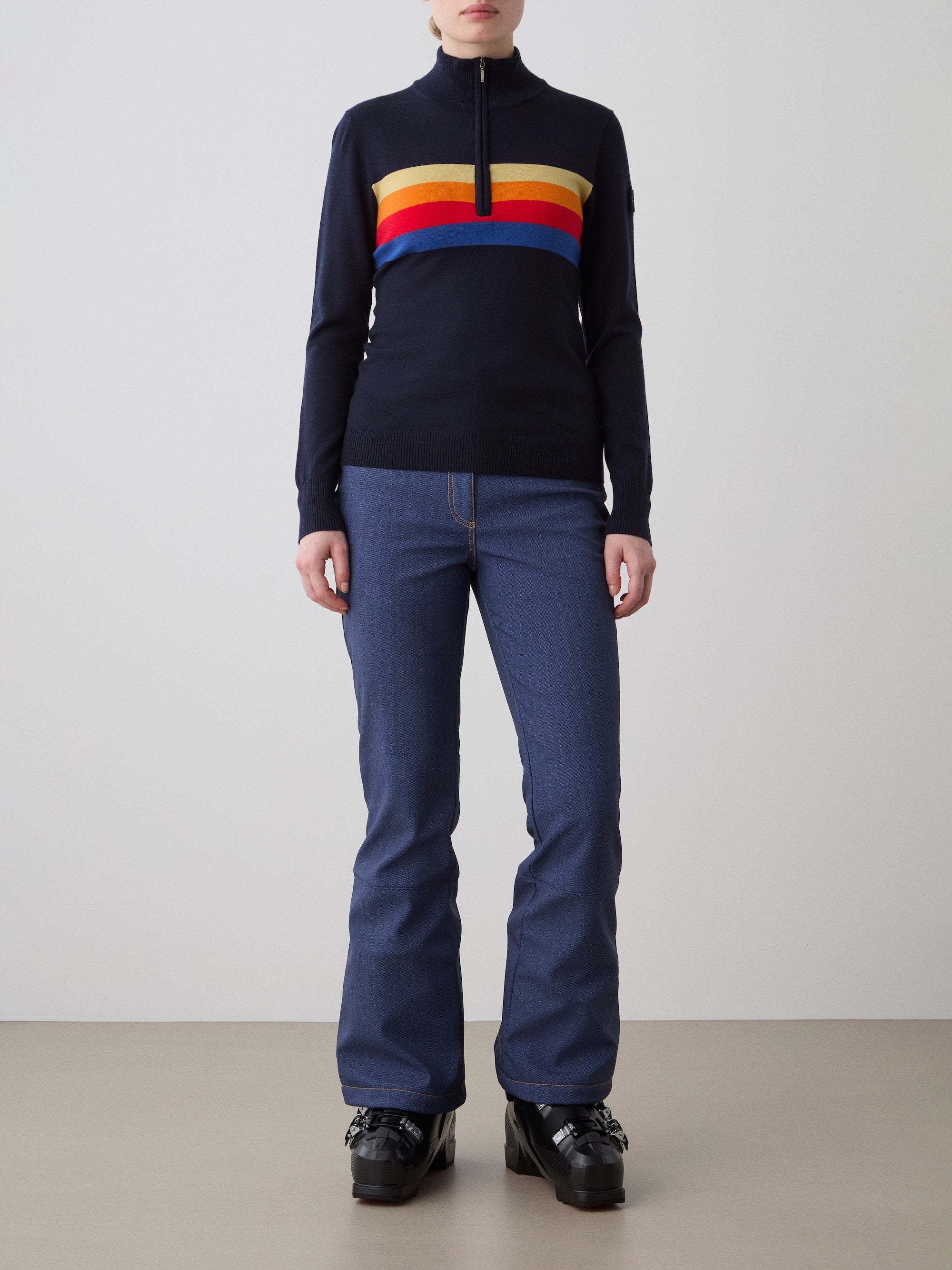 A person, face not visible, stands against a plain background wearing the We Norwegians FreeBird ZipUp women—a navy sweater with red, yellow, and orange stripes—paired with blue jeans and black platform shoes.