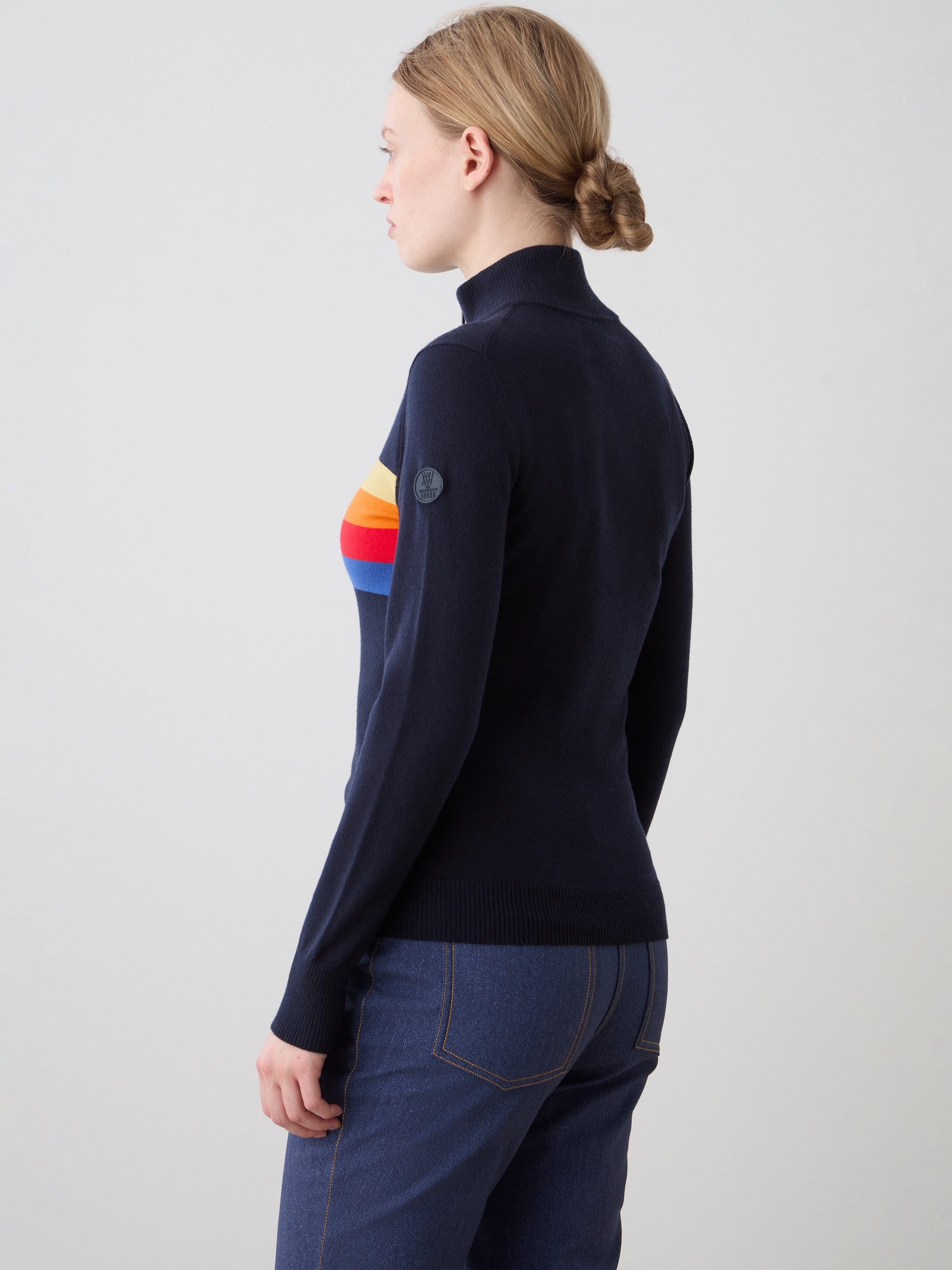 A woman with blond hair in a low bun models the We Norwegians FreeBird ZipUp, an alpine sweater with a colorful rainbow stripe across the chest, paired with dark blue jeans. She faces away against a plain light background.