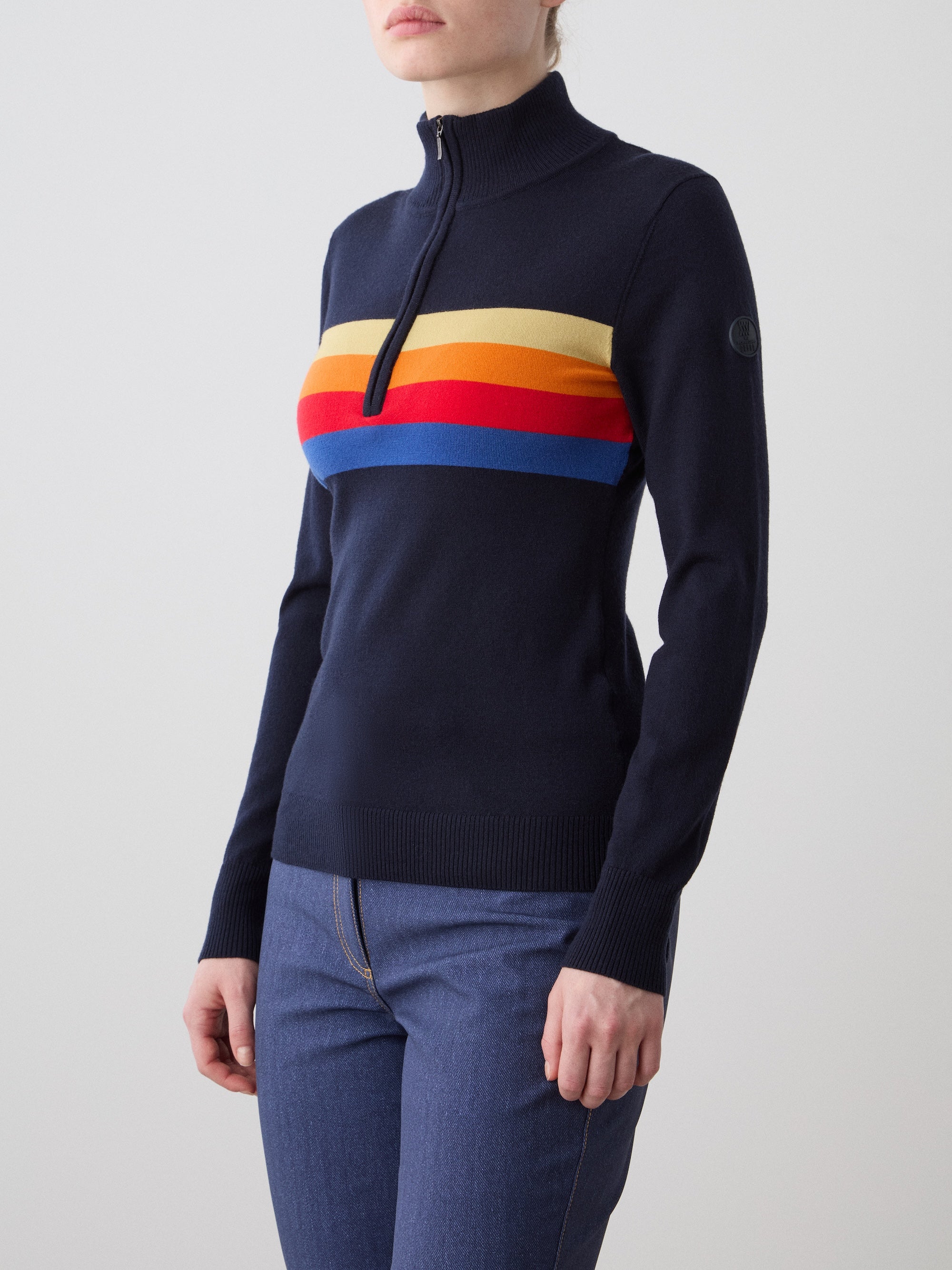A woman wears the We Norwegians FreeBird ZipUp in navy blue with yellow, orange, red, and blue chest stripes, styled with blue jeans, standing against a plain light background.