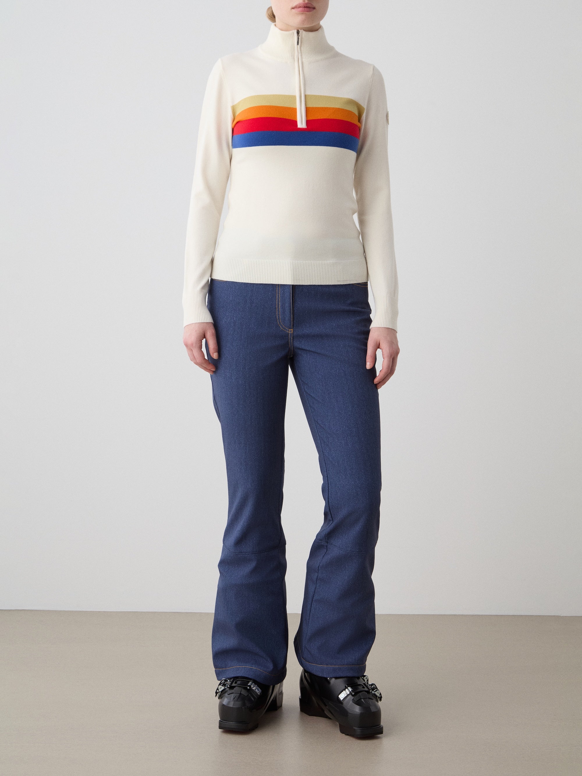 A person stands against a plain background wearing the We Norwegians FreeBird ZipUp women’s sweater with red, orange, and blue stripes, paired with blue jeans and black platform shoes. Their hands are relaxed at their sides.