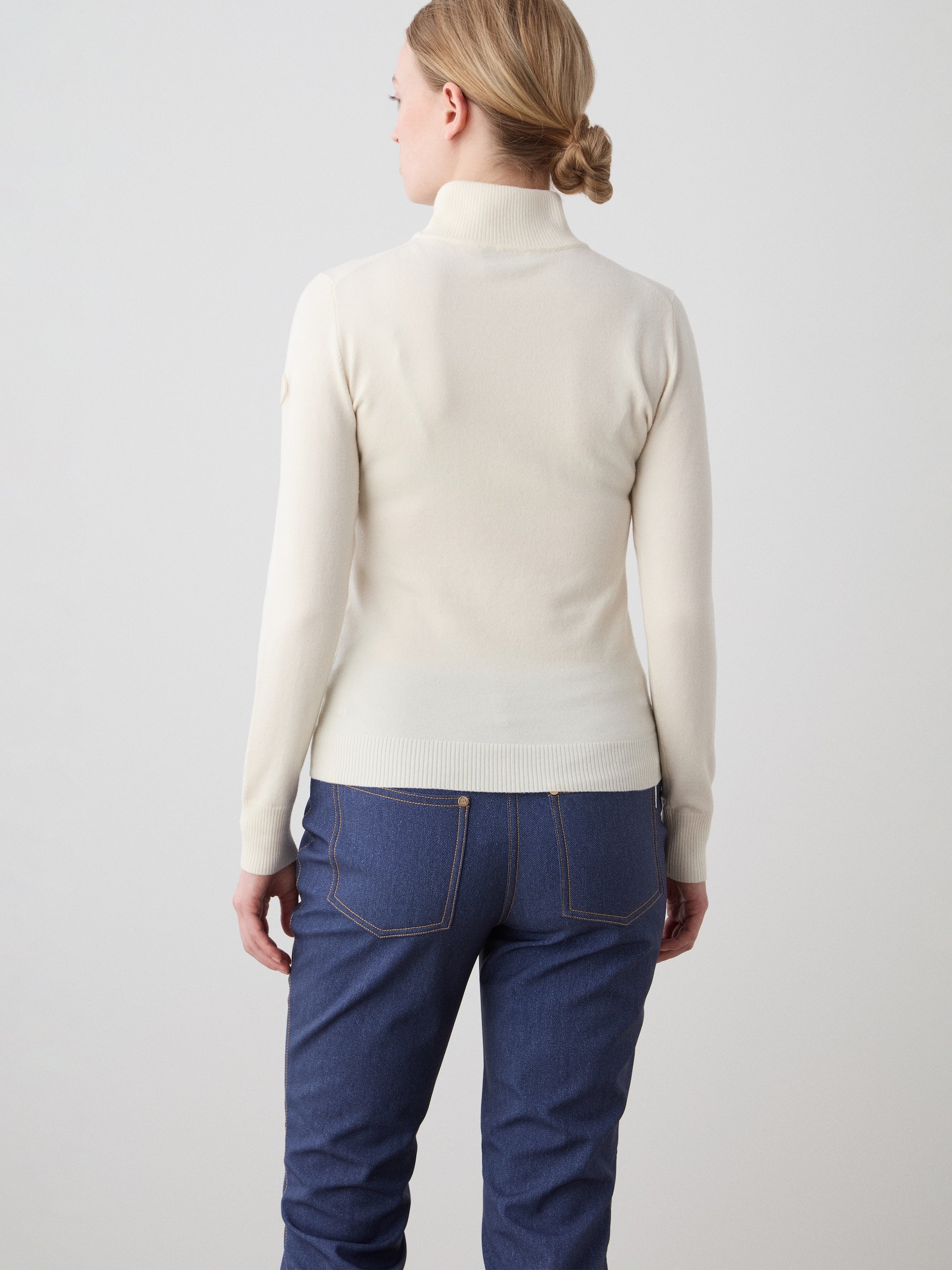 A woman with blonde hair in a low bun models the We Norwegians FreeBird ZipUp sweater, paired with blue jeans, standing with her back to the camera against a plain light grey background.