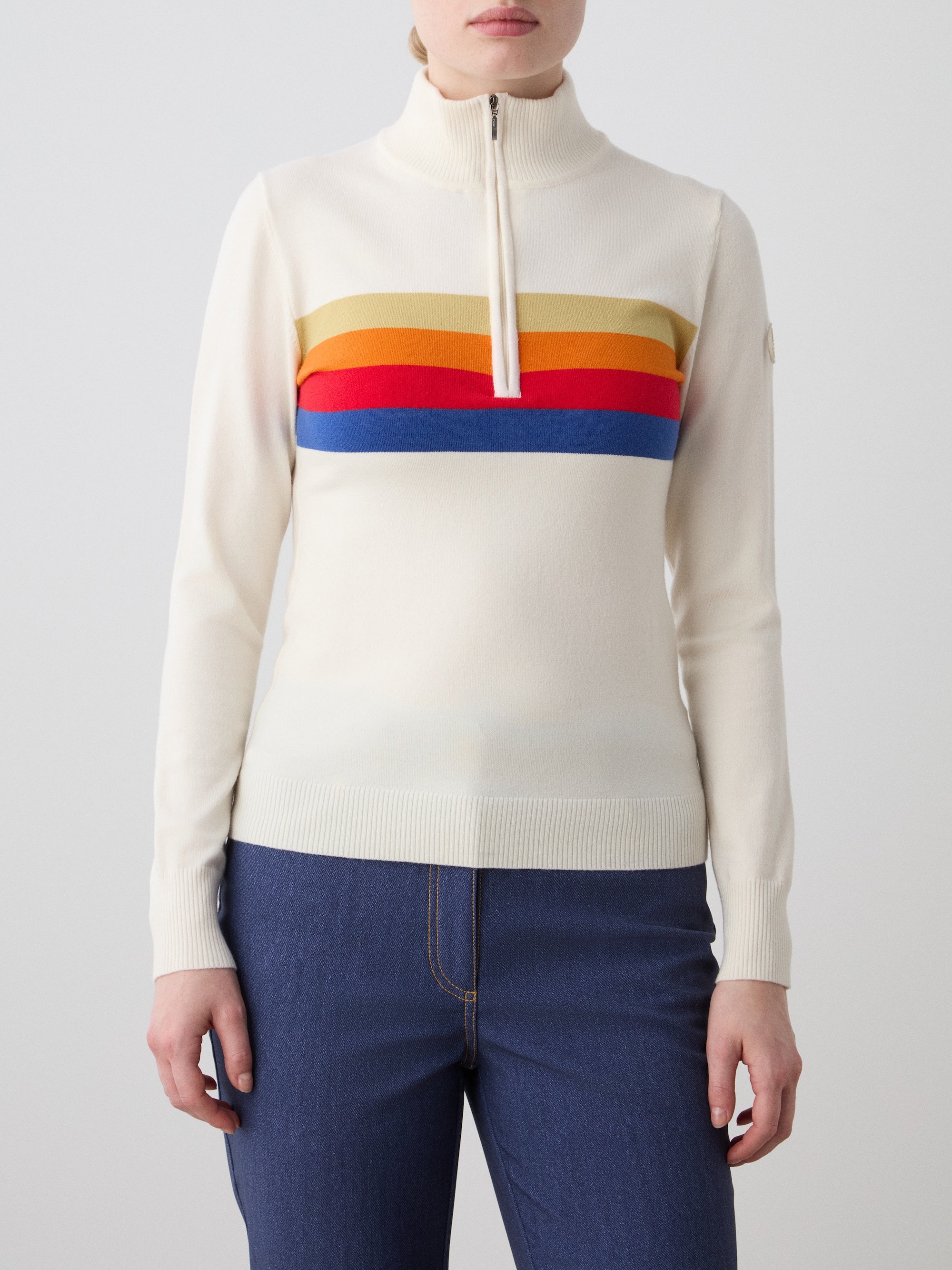 A person wears the We Norwegians FreeBird ZipUp women’s Merino wool sweater in cream with colorful chest stripes—yellow, brown, red, orange, and blue. This cozy long-sleeve pairs perfectly with blue jeans for a stylish look.