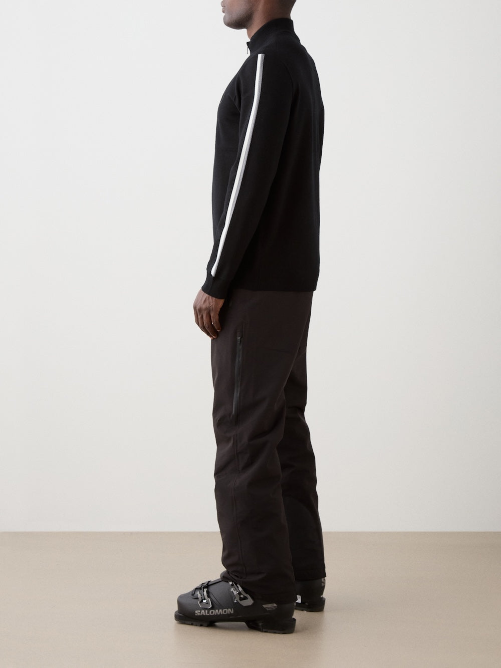 The model is wearing The We Norwegians FreeBird ZipUp men is a black long-sleeve ski sweater with a quarter-zip collar, white stripes on the shoulders and sleeves, and a subtle circular logo on the left chest.