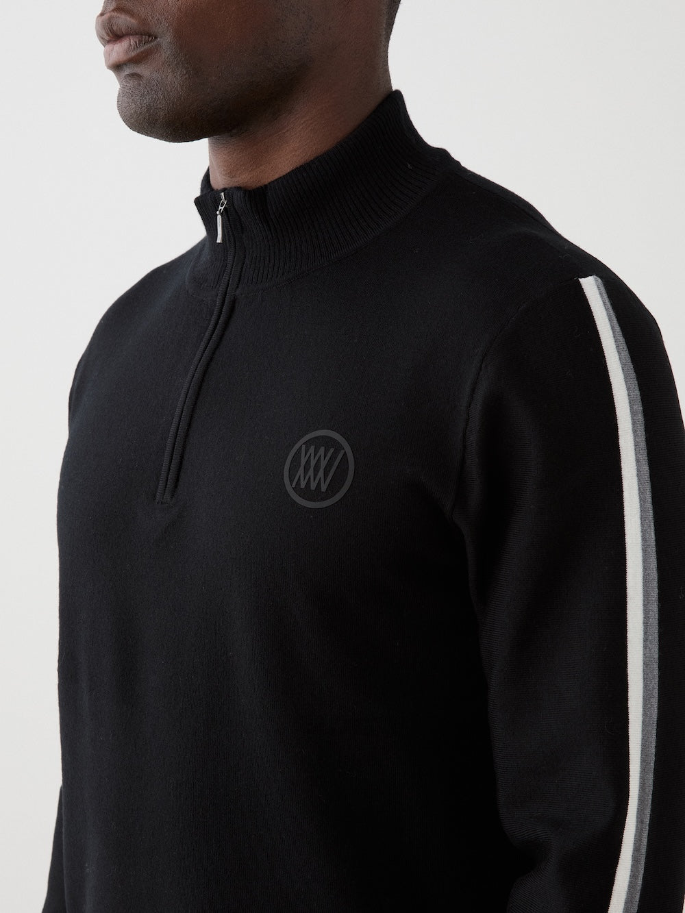 The model is wearing The We Norwegians FreeBird ZipUp men is a black long-sleeve ski sweater with a quarter-zip collar, white stripes on the shoulders and sleeves, and a subtle circular logo on the left chest.
