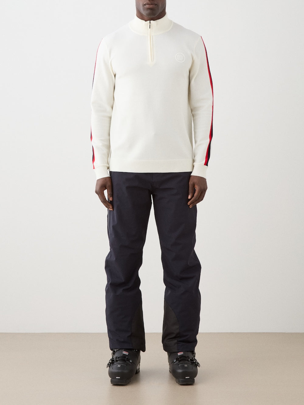 Model is wearing The We Norwegians FreeBird ZipUp men is a cream-colored, lightweight quarter-zip sweater with red and black sleeve stripes and a white circular chest logo—a modern update of the classic ski sweater.