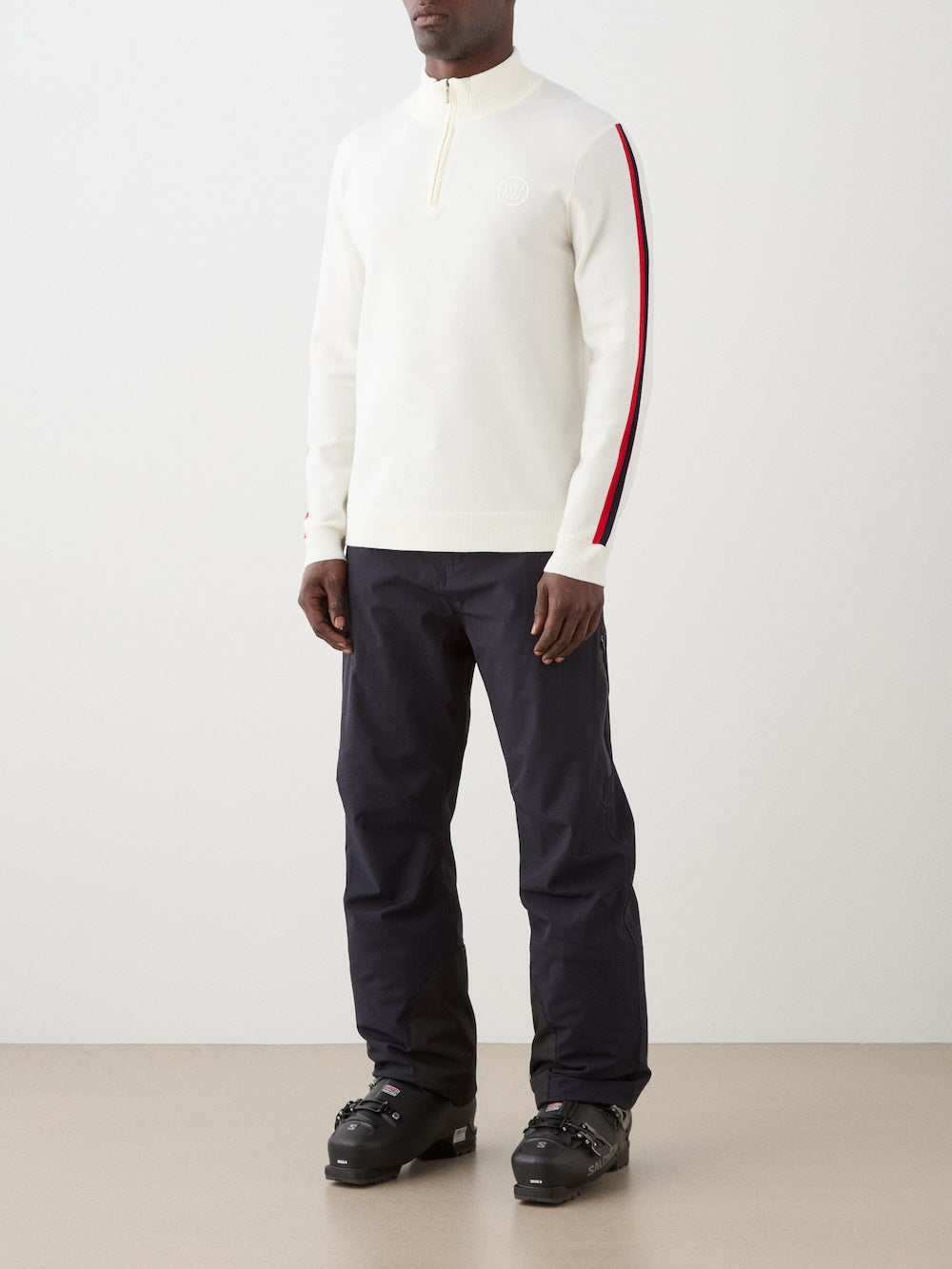 Model is wearing The We Norwegians FreeBird ZipUp men is a cream-colored, lightweight quarter-zip sweater with red and black sleeve stripes and a white circular chest logo—a modern update of the classic ski sweater.