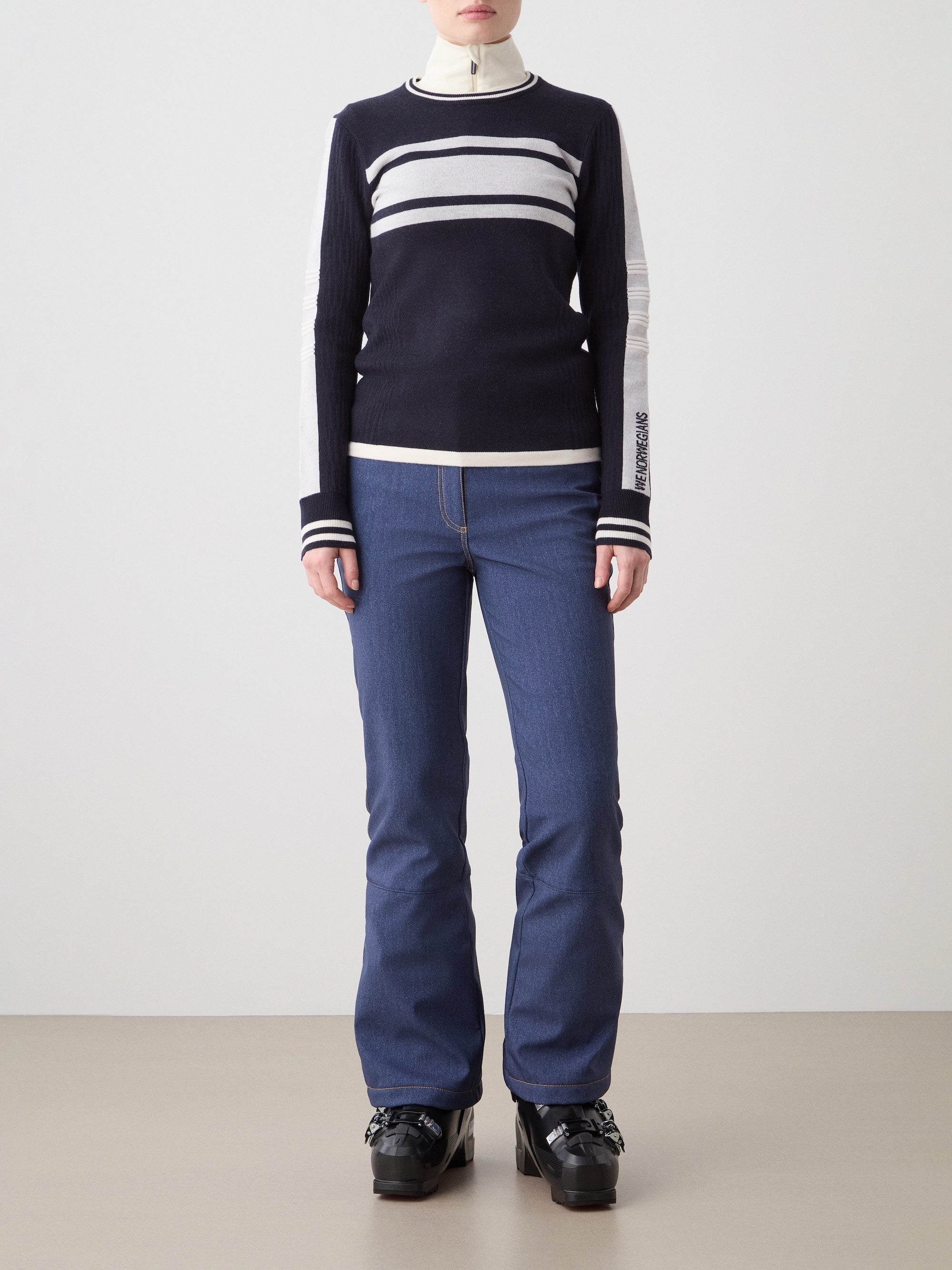 A woman stands against a plain background wearing the We Norwegians Rocky Ski Sweater in navy, white, and black with a retro ski vibe, paired with blue jeans and chunky black shoes. Her hands are by her sides.