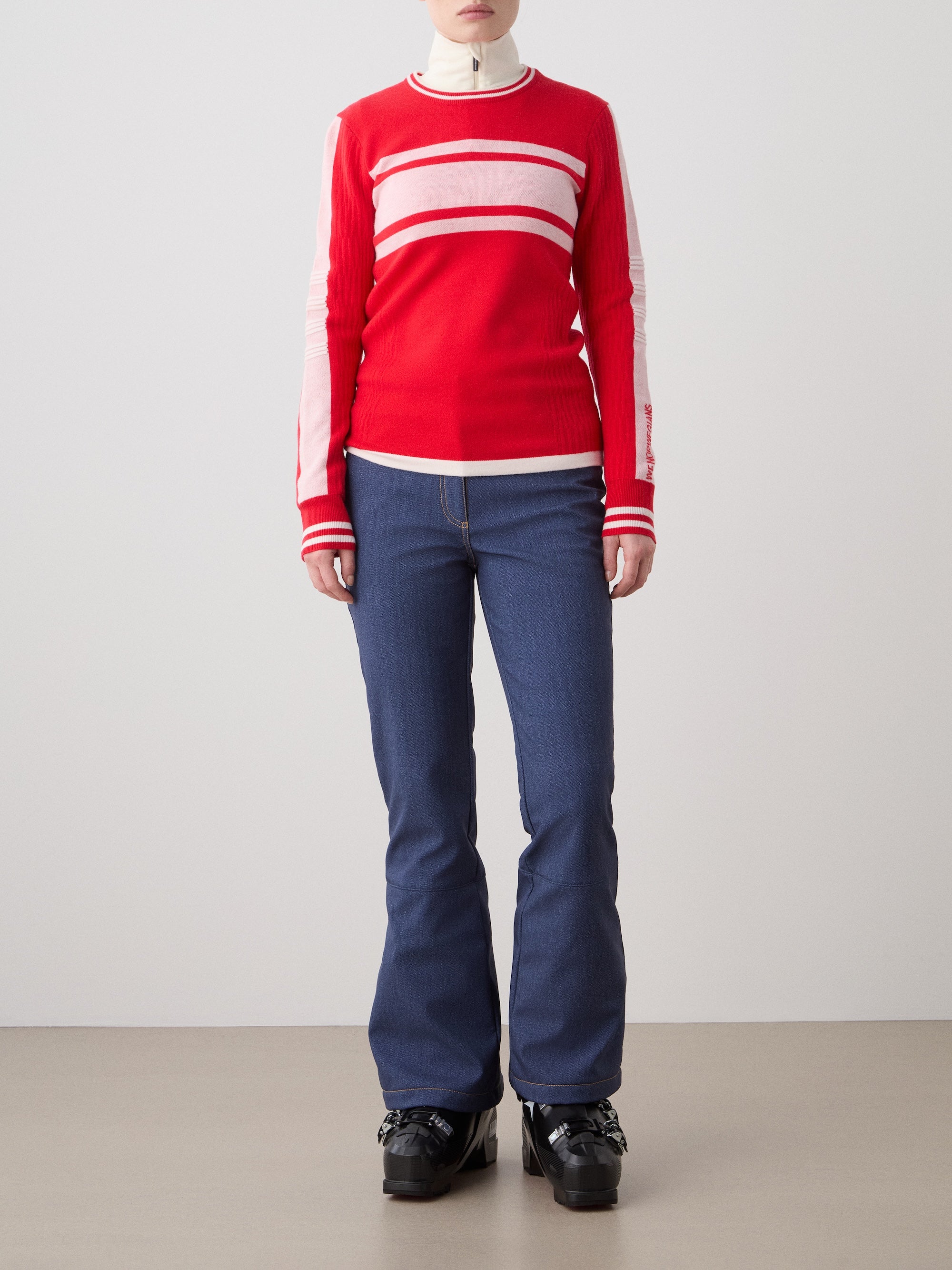 A person stands against a plain background wearing the Rocky Ski Sweater for women by We Norwegians, paired with blue jeans and black platform shoes. Their hands are at their sides and their face is not visible.