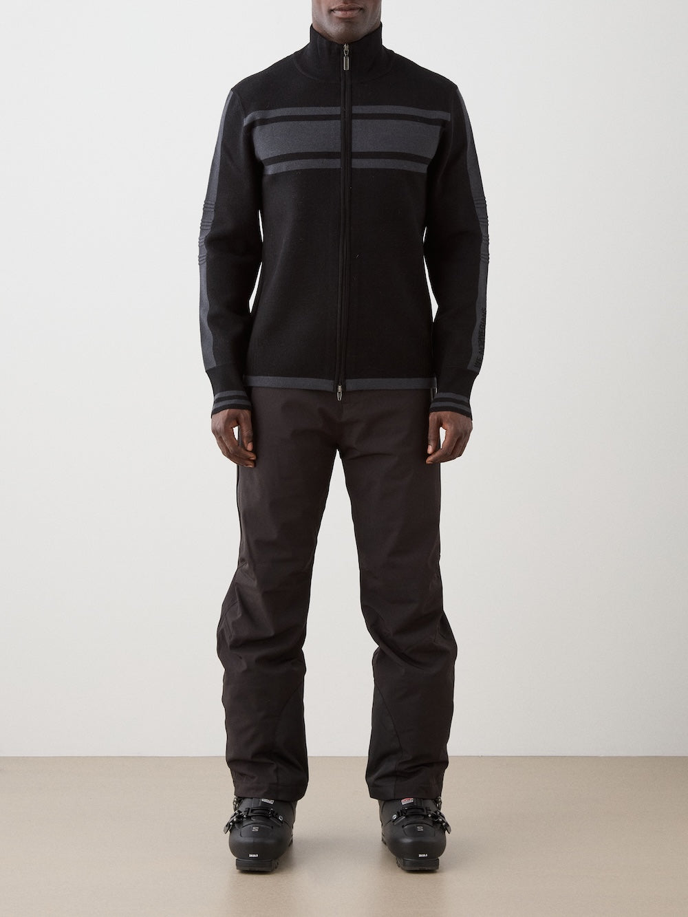 A person stands facing forward, wearing the We Norwegians Rocky FullZip men’s retro racing knit jacket with gray stripes, dark pants, and black ski boots, against a plain, neutral background.