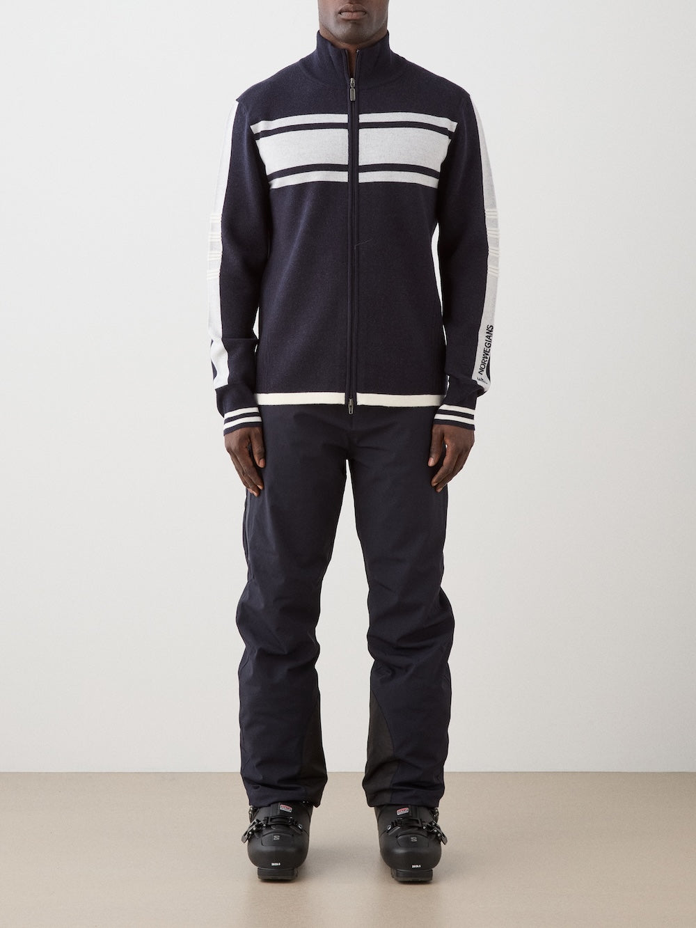 A person is wearing the We Norwegians Rocky FullZip men navy retro racing knit zip-up jacket with white stripes, paired with black pants and black ski boots, standing against a plain light background.