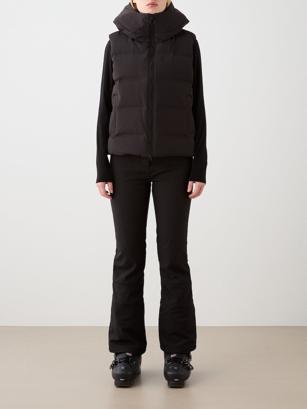 A woman faces forward wearing the black We Norwegians Aurora Vest—crafted as a responsible down vest—layered over a black long-sleeve top with black pants and ski boots. The minimalist scene reflects Scandinavian style against a plain white wall and beige floor.