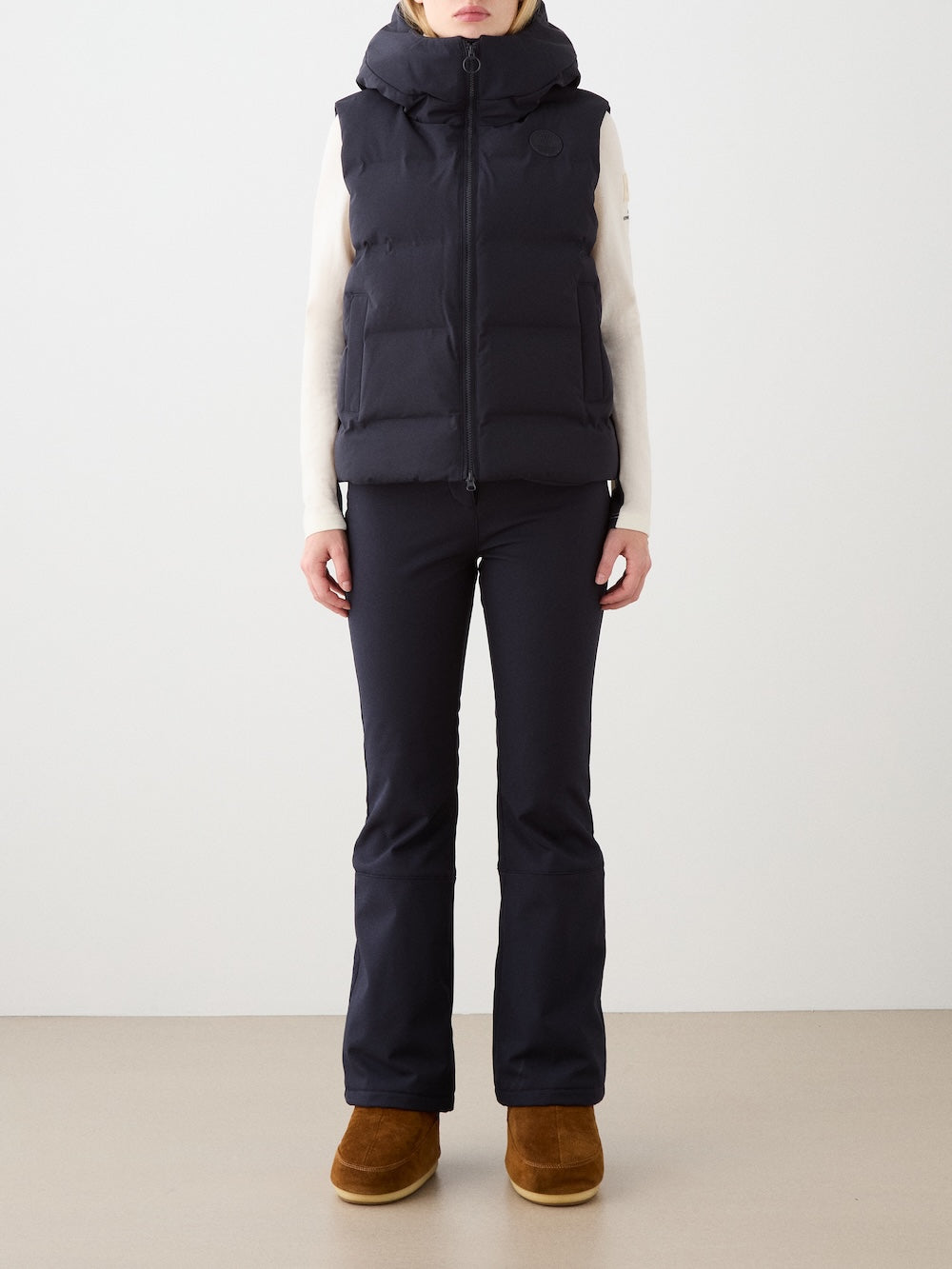 A woman stands against a plain background, showcasing the We Norwegians Aurora Vest women—a black, responsibly-made down vest—styled over a light long-sleeve top, black pants, and brown suede boots for a clean Scandinavian look.