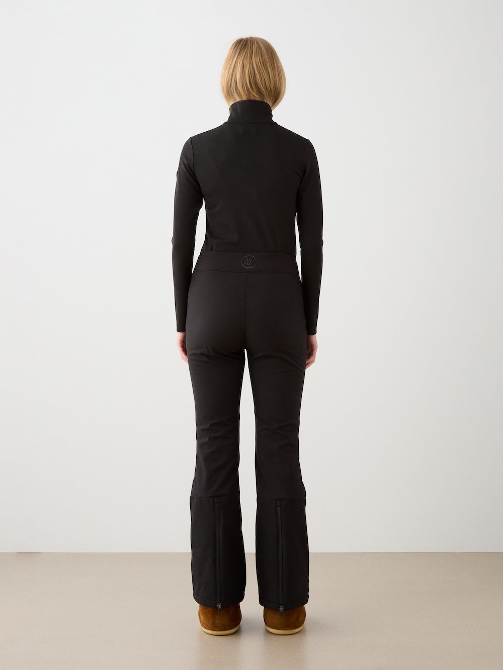 A woman, seen from behind, wears the We Norwegians Bibi ZipUp women in black with matching flared pants and brown boots, evoking vintage ski style against a light background.