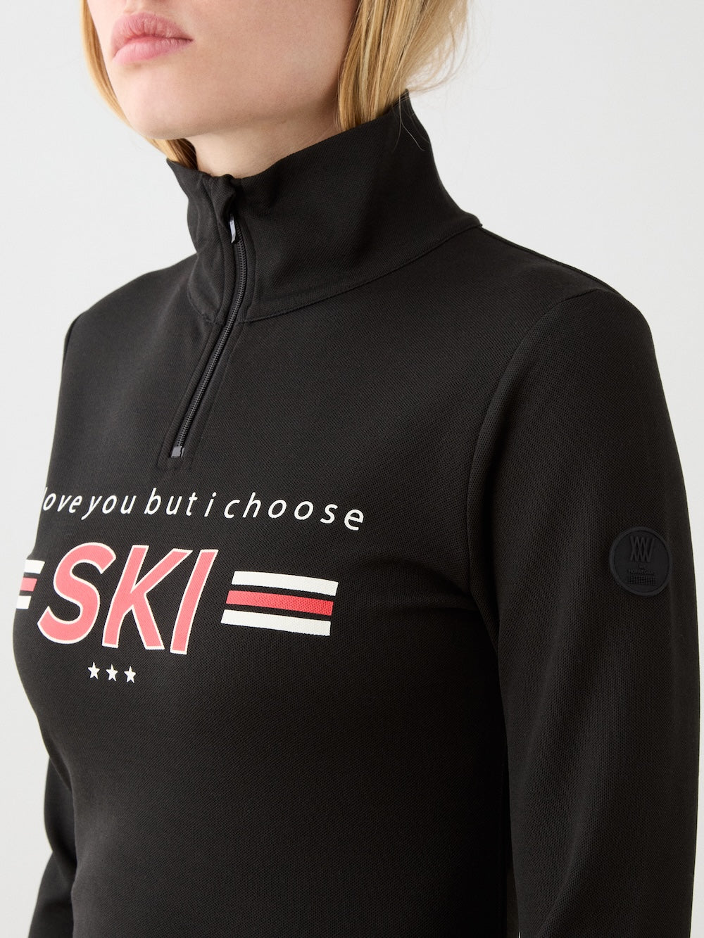 A woman models the We Norwegians Bibi ZipUp women—a vintage-inspired black ski top with red and white chest stripes and "I love you but I choose SKI"—against a plain light background.
