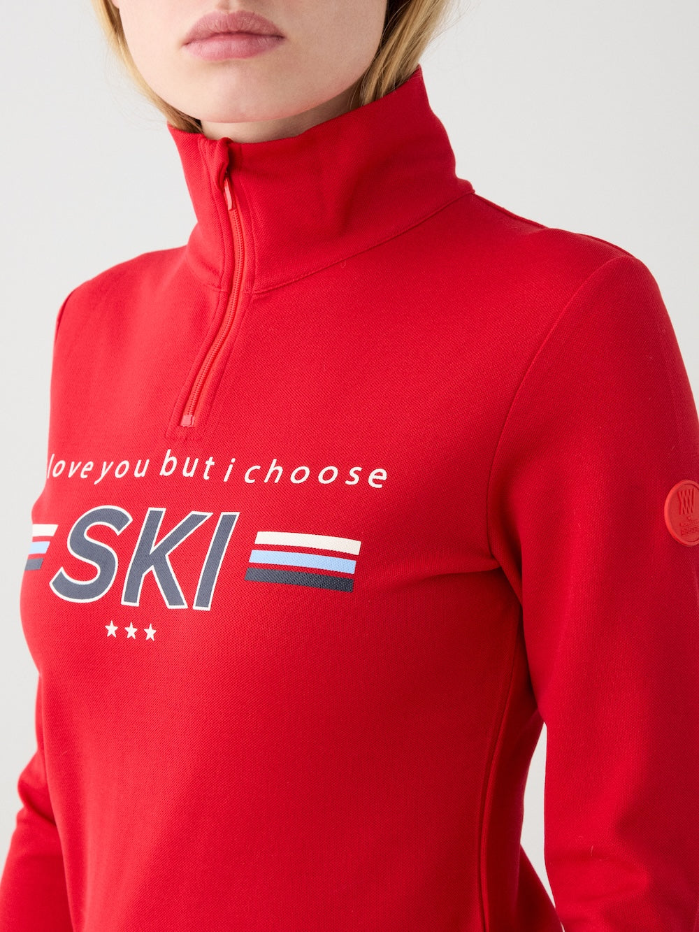 A woman wears the We Norwegians Bibi ZipUp in red, a vintage-inspired quarter-zip ski top with "I love you but I choose SKI," plus decorative stripes and stars. Only her upper body and lower face are shown.