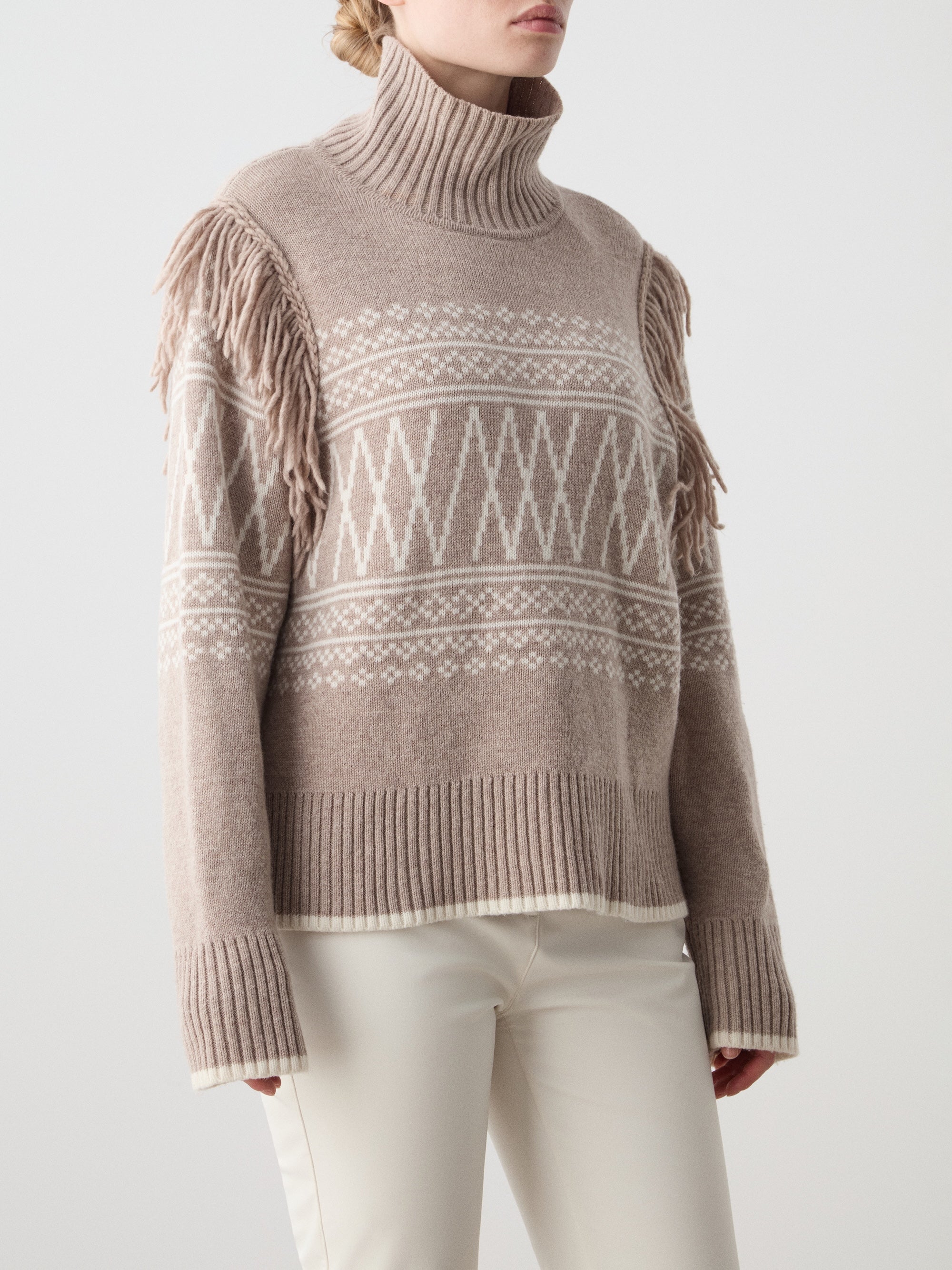 A woman models the We Norwegians Setesdal Sweater for women—beige with white patterns and fringe at the shoulders—paired with white pants, set against a simple light background.