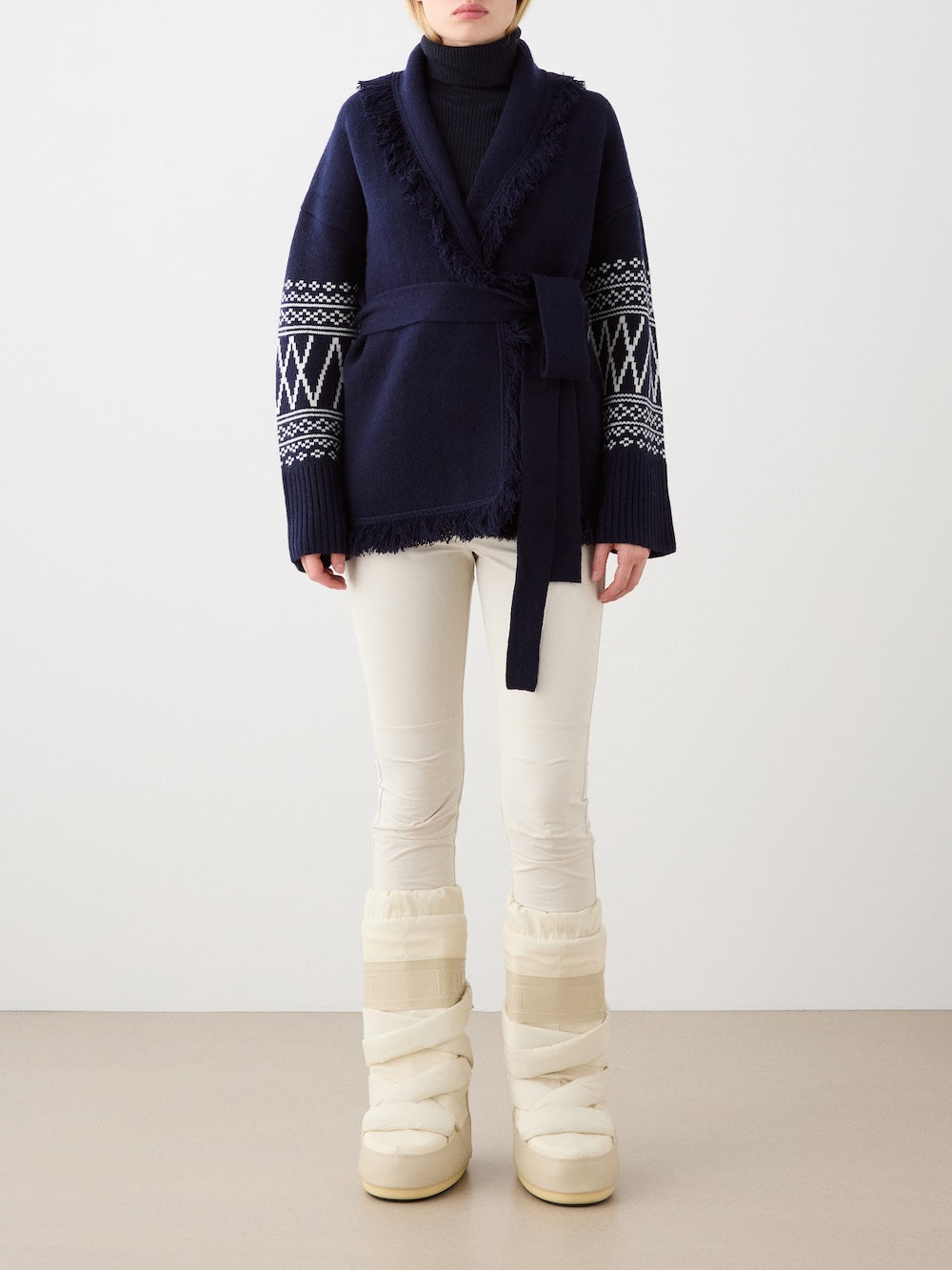 A woman wears the We Norwegians Setesdal Cardigan in navy blue with white sleeve patterns, paired with cream pants and beige puffer snow boots, standing against a plain, light background.