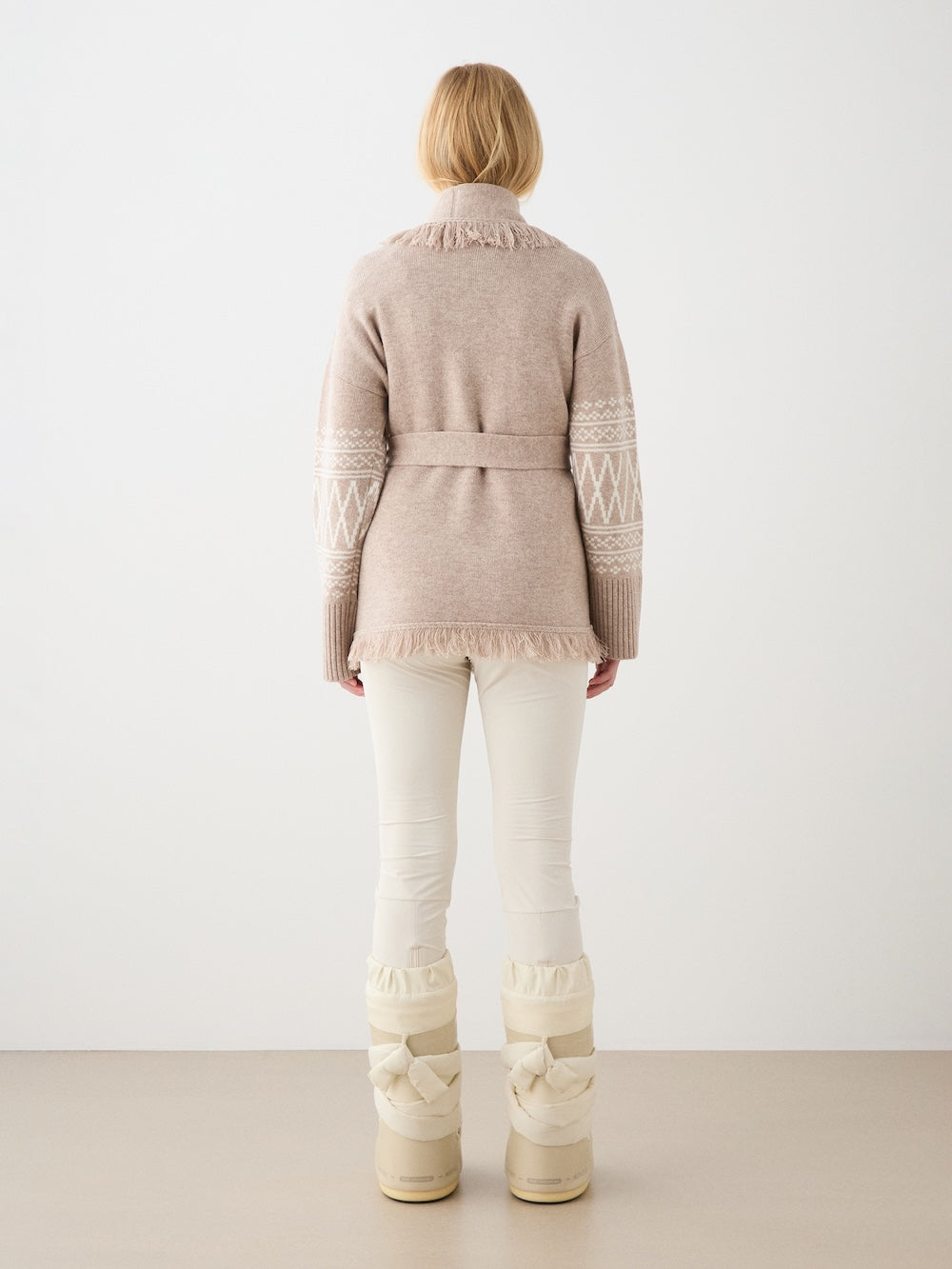A person with blonde hair, seen from behind, wears the We Norwegians Setesdal Cardigan for women with ivory pants and cream snow boots, standing against a plain neutral background.