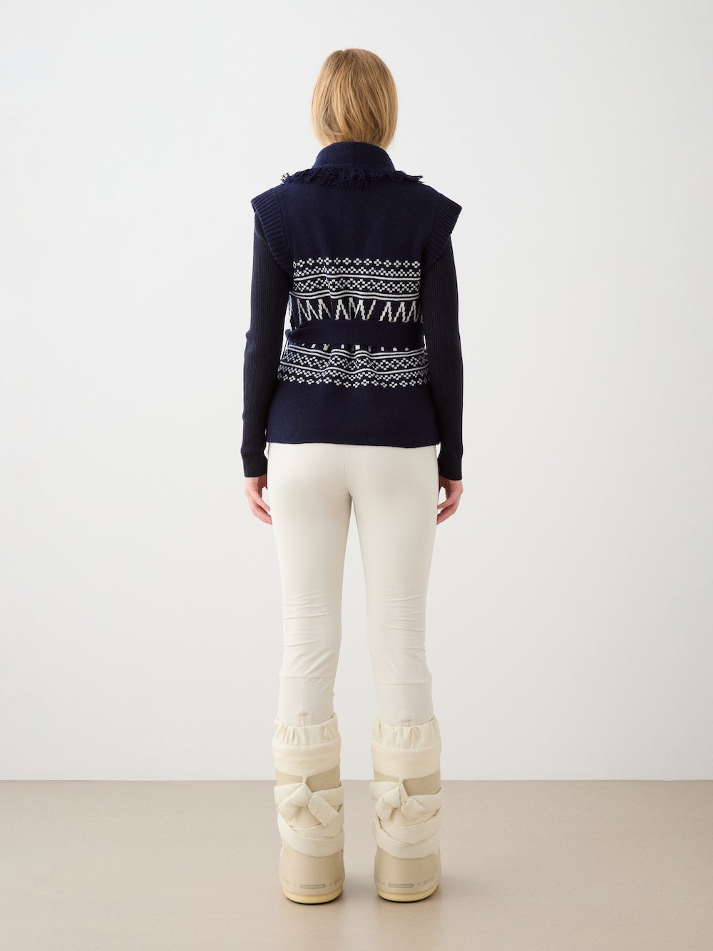 A person with light brown hair stands facing away, wearing the We Norwegians Setesdal Vest for women, paired with cream pants and cream quilted winter boots, against a plain white background.
