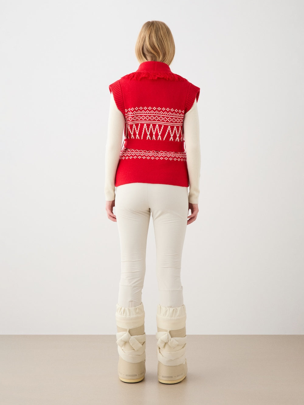 A person stands facing away in a We Norwegians Setesdal Vest women—red Norwegian knitwear with white geometric patterns—layered over a cream long-sleeve top, paired with cream pants and beige padded winter boots in a minimalist setting.