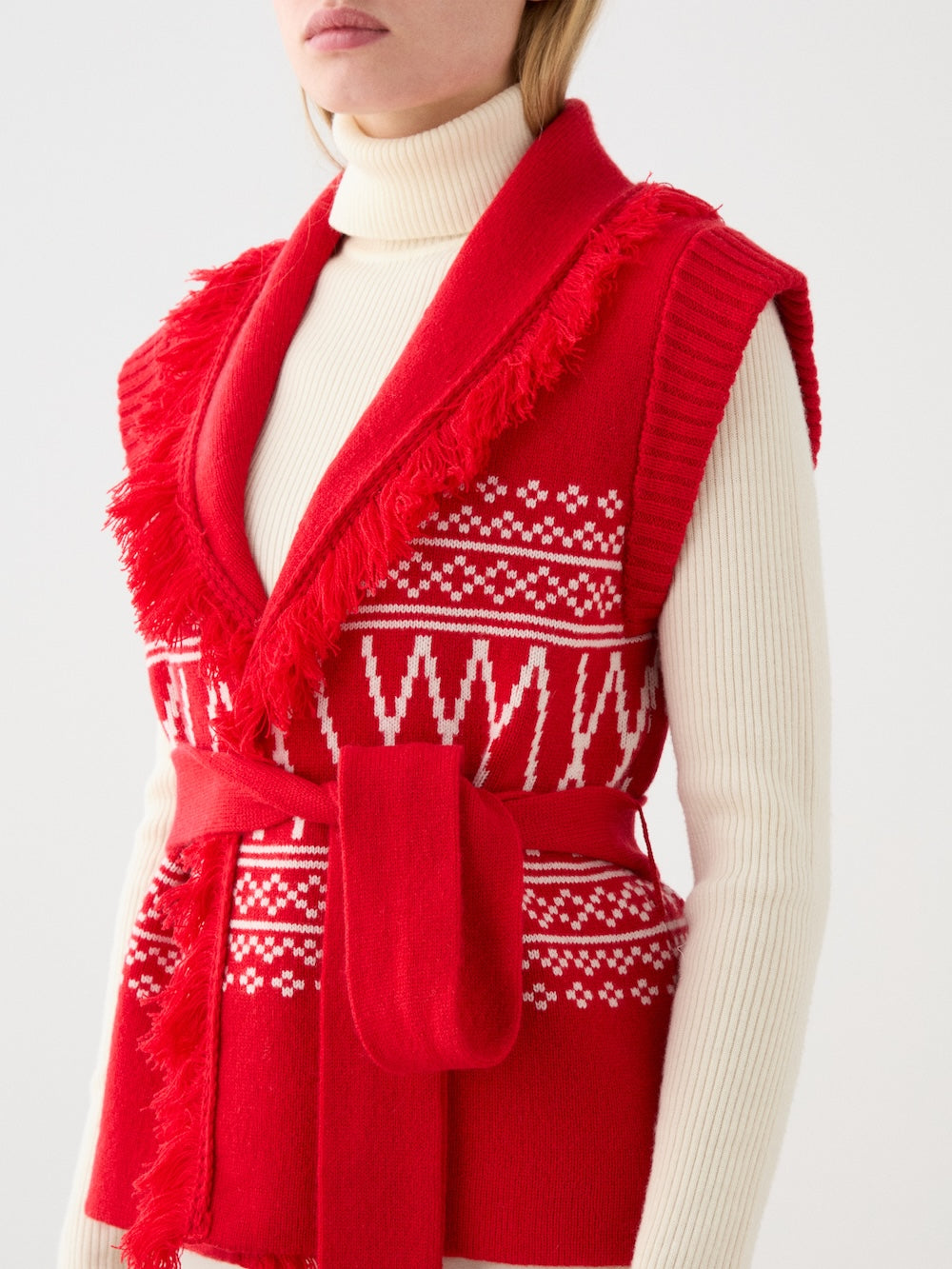 A person wears a cream turtleneck and the We Norwegians Setesdal Vest for women, which is red with white geometric patterns, a matching waist belt, and fringe trim. The image is cropped at the shoulders and mid-thigh.