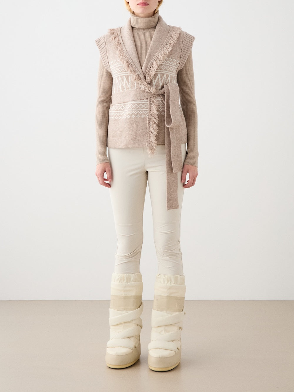 A person stands against a plain background wearing a beige turtleneck sweater, white fitted pants, chunky cream snow boots, and the Setesdal Vest by We Norwegians. Their face is not visible.
