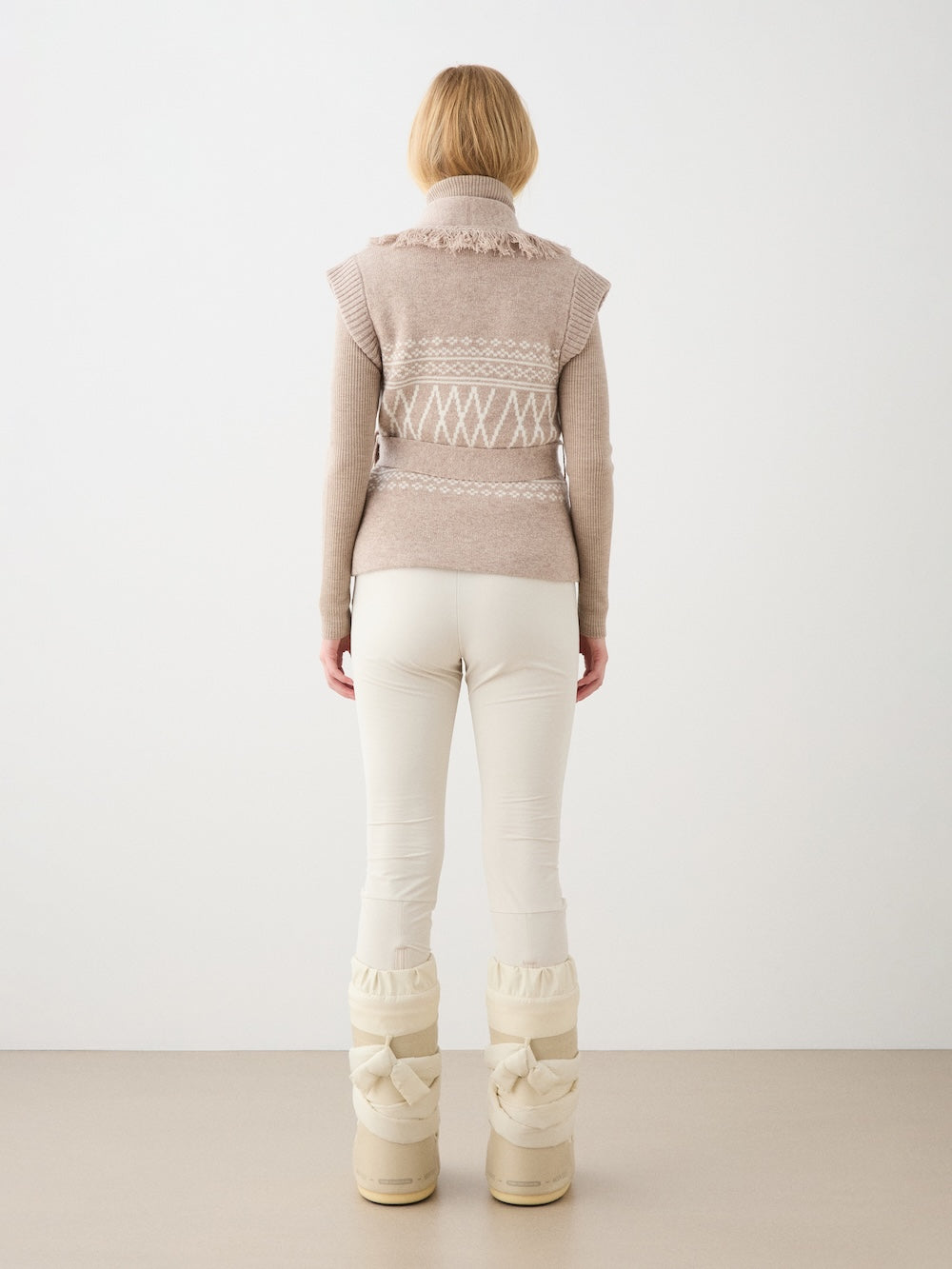 A person with light hair faces away, wearing the We Norwegians Setesdal Vest for women—a beige Norwegian knitwear vest with white patterns—over a long-sleeve top, paired with white pants and beige snow boots, against a plain white background.