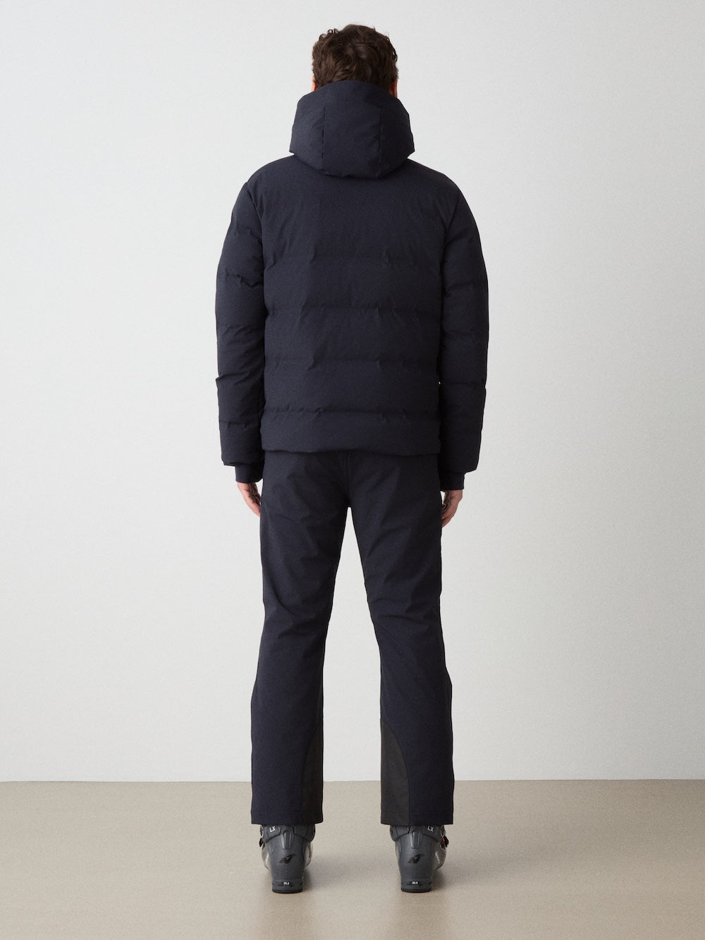 A person faces away, wearing the We Norwegians Nordlys Ski Jacket for men—black, waterproof, with a hood—paired with black pants and gray ski boots, set against a plain light background.