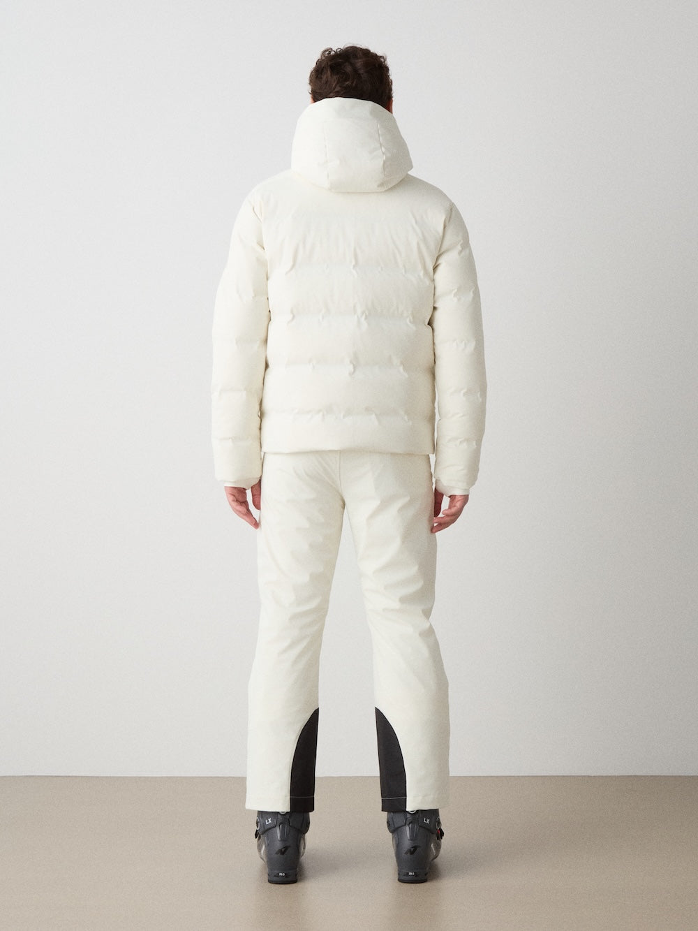 A person faces away against a plain backdrop, wearing the We Norwegians Nordlys Ski Jacket for men in white with a hood, matching pants featuring black patches on the lower legs, and dark snow boots.