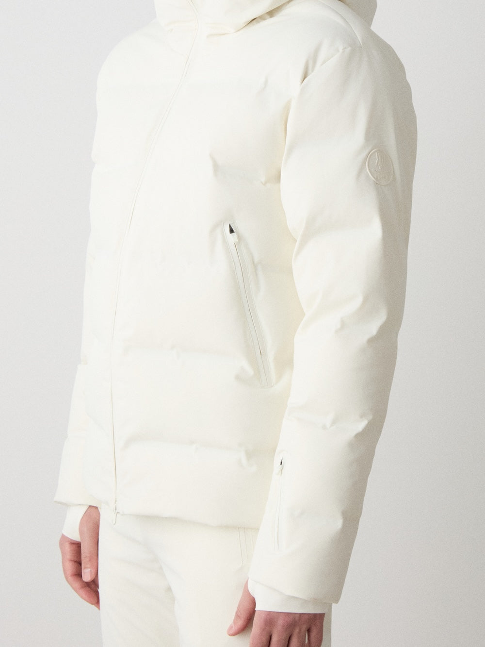 A person models the We Norwegians Nordlys Ski Jacket men in off-white, filled with responsible down and designed with a hood and zippered pockets, paired with matching pants. Only the torso and part of their arms are visible against a plain light background.