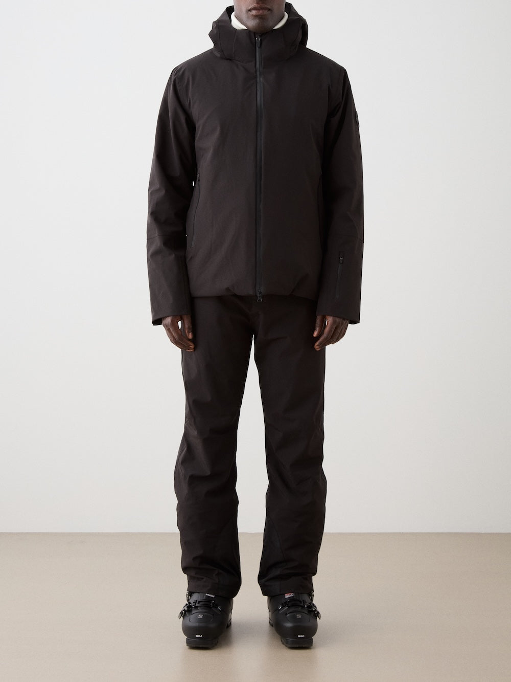 A man stands facing forward in a dark We Norwegians Hammerfest Ski Jacket and matching pants with black ski boots, set against a plain light background.