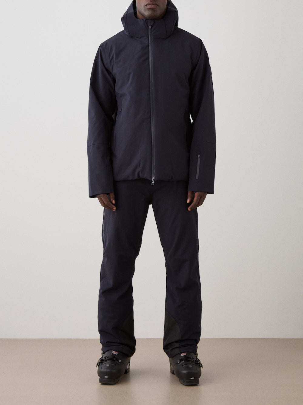 A person faces forward, wearing the black We Norwegians Hammerfest Ski Jacket for men with matching black pants and ski boots, set against a plain, light background.
