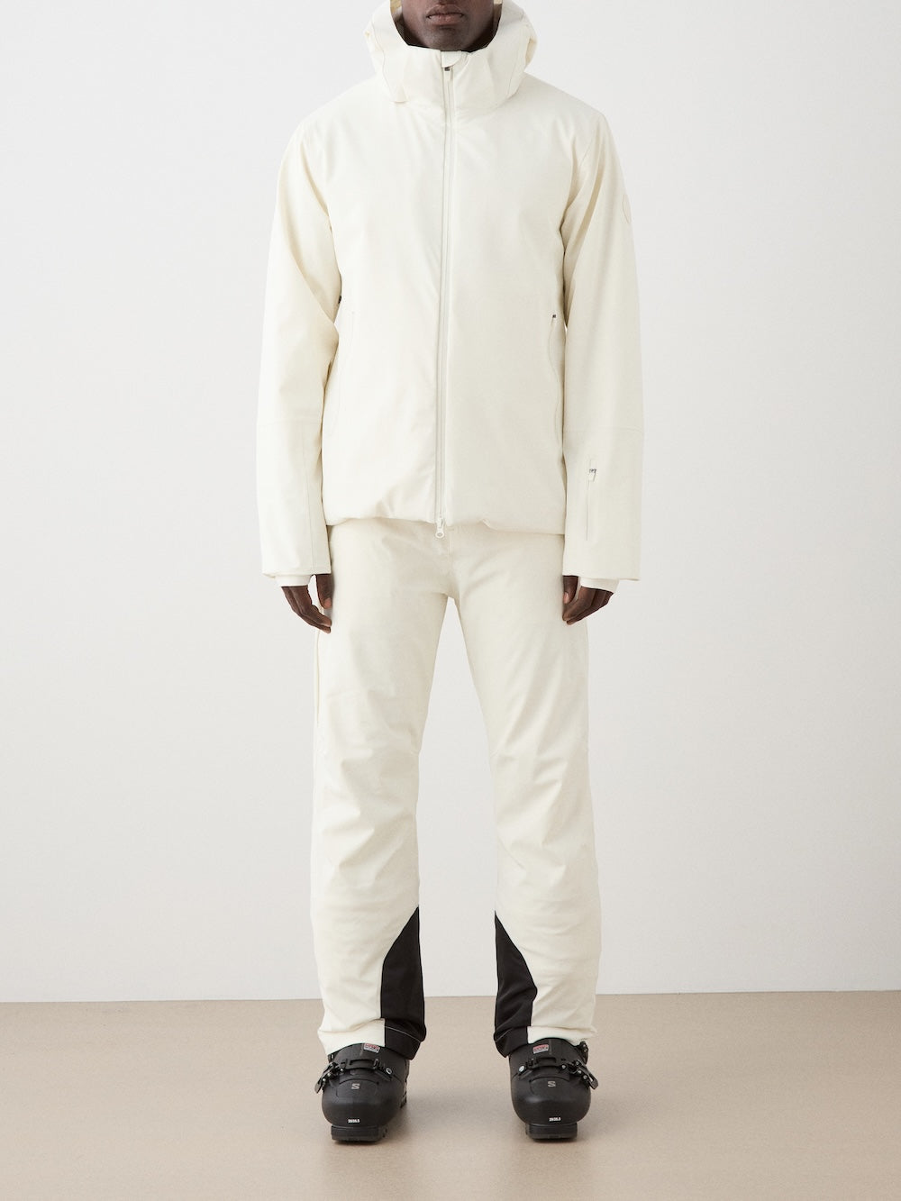 A person stands against a plain background wearing the We Norwegians Hammerfest Ski Jacket men and matching pants with black ski boots. The minimalist outfit features clean lines, 4-way stretch fabric, and a sleek waterproof design.