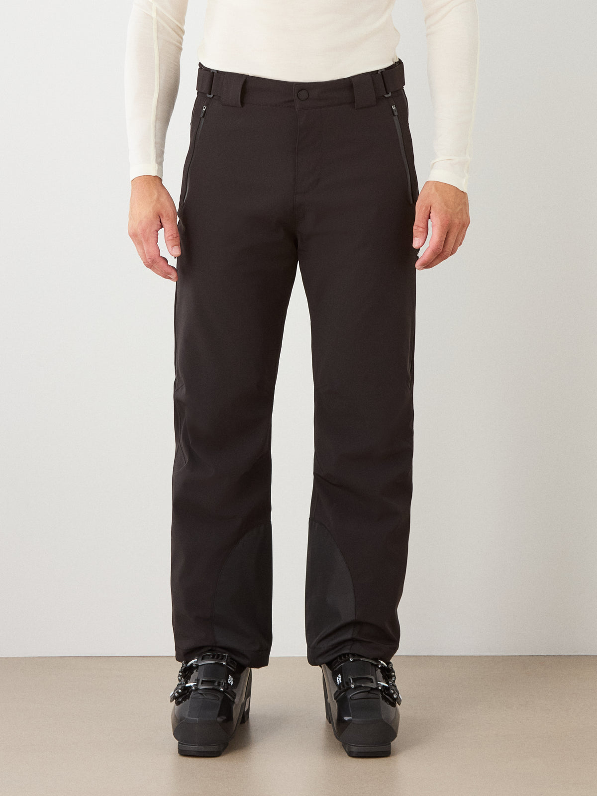 A person wearing a white long-sleeve base layer and We Norwegians Hammerfest Ski Pant for men in black, paired with black ski boots, stands against a plain light gray background.