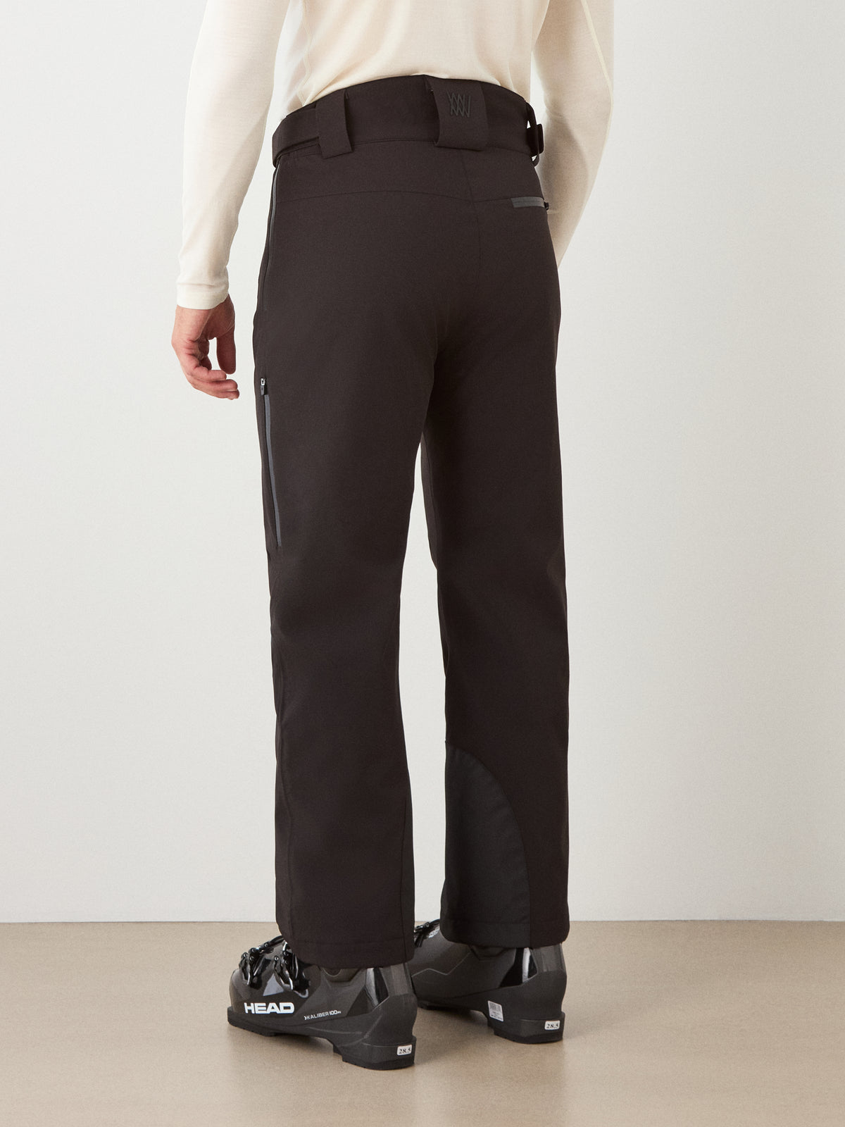 A person stands indoors on a plain background, facing away and wearing the We Norwegians Hammerfest Ski Pant for men—black ski pants featuring a side zipper pocket and reinforced ankle patches. The outfit is completed with a light long-sleeve shirt and black HEAD ski boots. These waterproof pants combine style and durability, making them an excellent choice for those seeking high-quality men’s ski apparel.