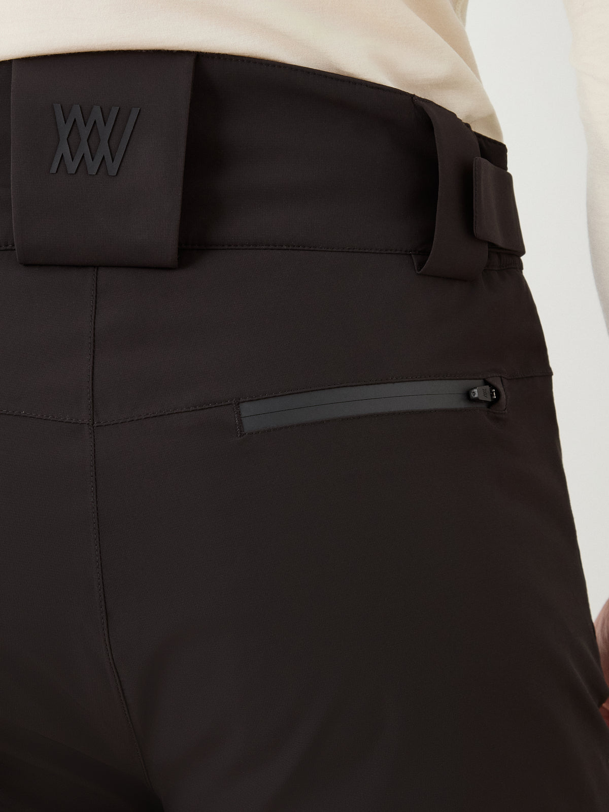 Close-up of the Hammerfest Ski Pant men by We Norwegians, shown in black. The ski pants feature a prominent, embossed “XXV” logo on the waistband and a horizontal, water-resistant zippered pocket on the hip. Made with wool insulation and waterproof fabric, the pants are styled with a cream-colored top against a light gray background.