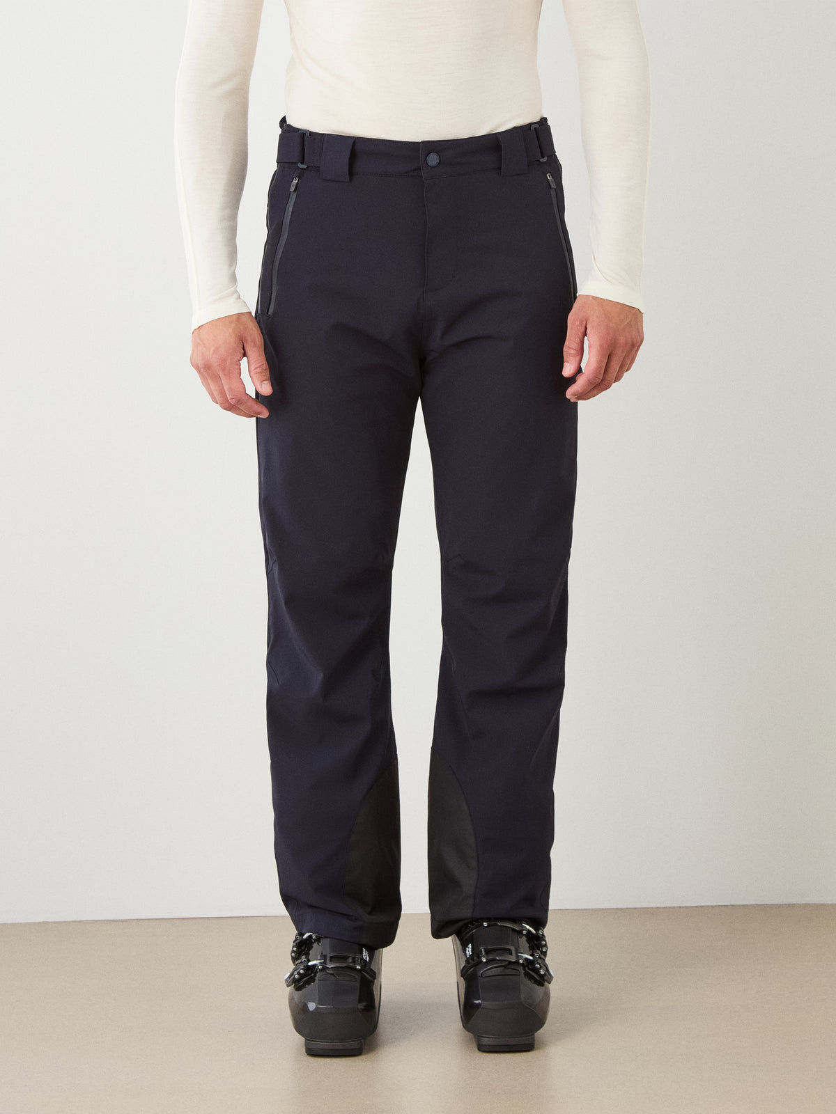 A person wearing We Norwegians Hammerfest Ski Pant men in black and black boots stands against a plain wall, shown from the shoulders down. The long-sleeved shirt is light-colored, and their hands are relaxed by their sides.
