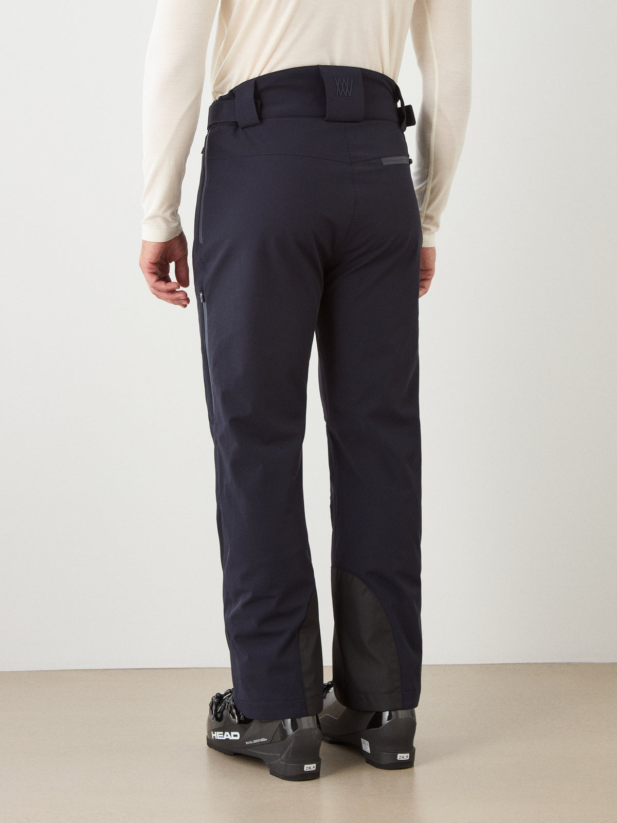 A person wearing We Norwegians Hammerfest Ski Pant for men in black, black ski boots, and a long-sleeve white top, standing indoors and facing away from the camera.