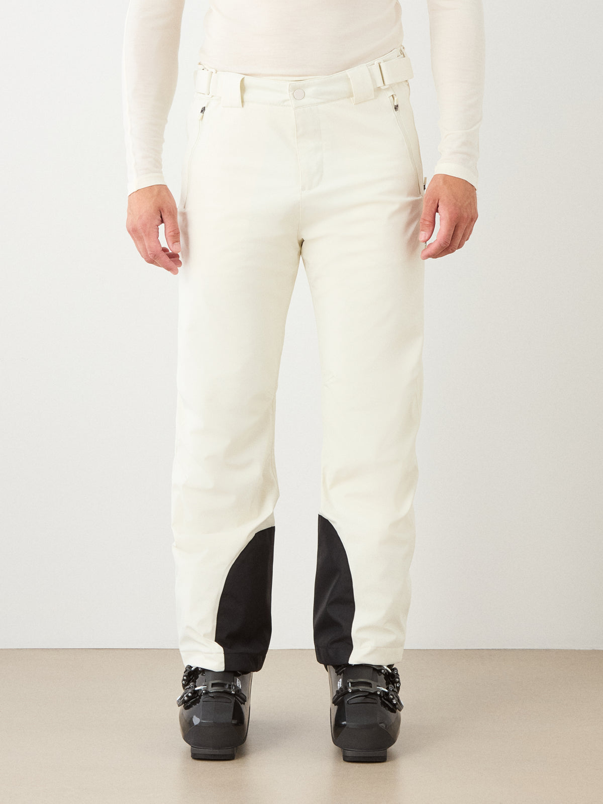 A person wearing the We Norwegians Hammerfest Ski Pant men—white, waterproof ski pants featuring black patches near the ankles—paired with a matching white long-sleeve top and black ski boots, stands against a plain background.