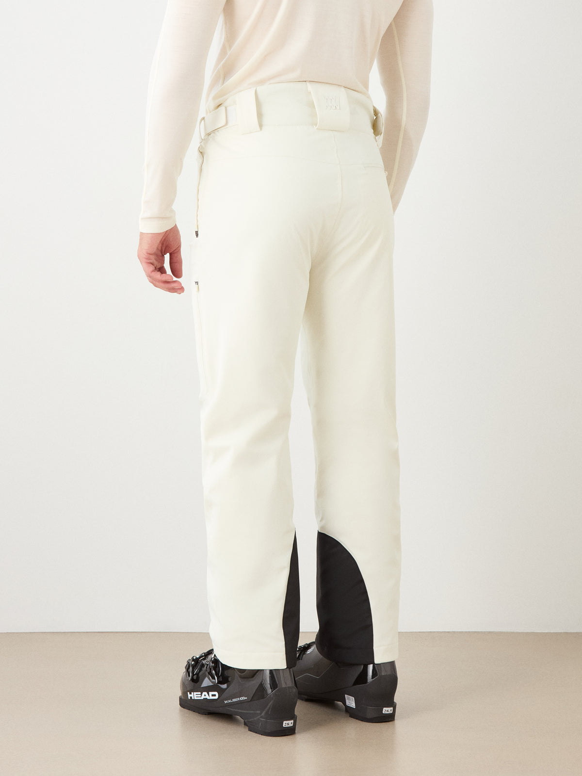 A person wearing We Norwegians’ Hammerfest Ski Pant men in ivory, paired with a beige top and black ski boots, stands facing away from the camera against a plain background.
