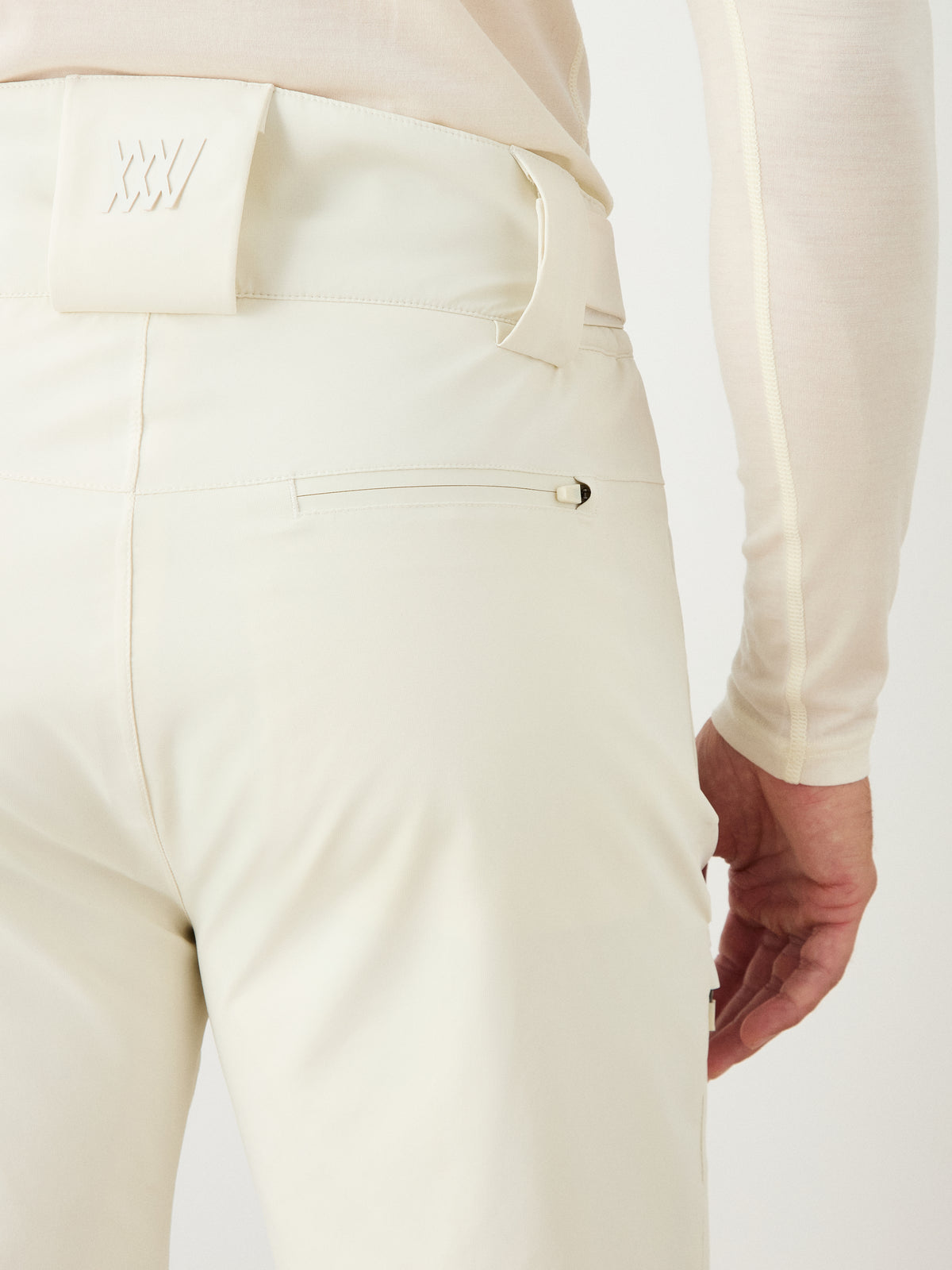 A close-up of a person wearing the We Norwegians Hammerfest Ski Pant for men in a cream color, paired with a matching long-sleeve shirt, highlighting the back pocket, belt loop, and a waistband patch embossed with “XXV.”.