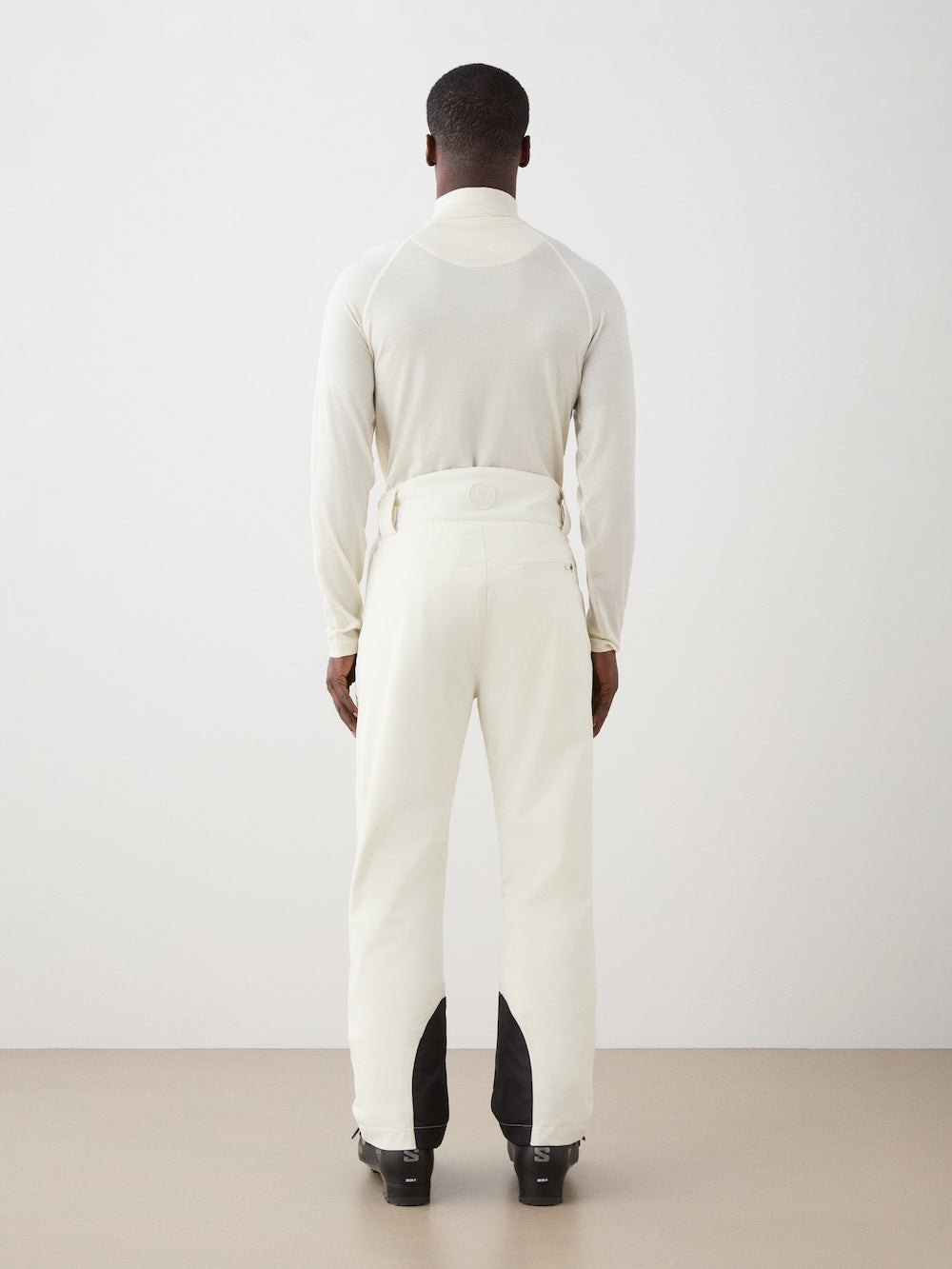 A man, seen from behind in a minimal, neutral room, wears an off-white long-sleeve shirt and We Norwegians Hammerfest Ski Pant men with black patches at the bottom.