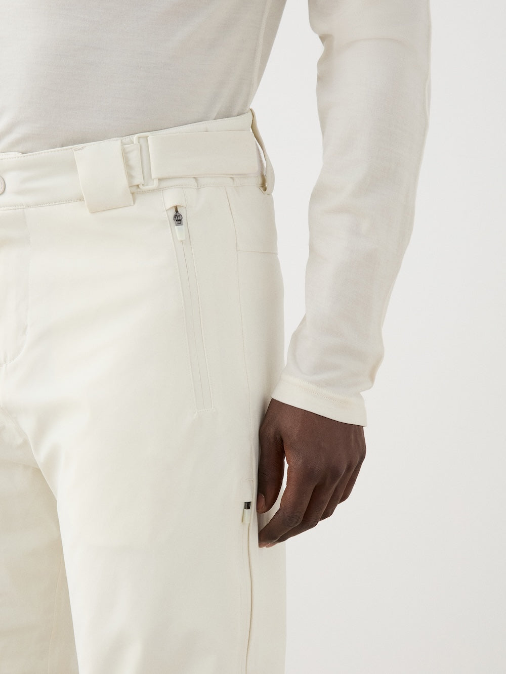 A person wears We Norwegians Hammerfest Ski Pant men in off-white wool with a belt and a matching long-sleeve top. The shot highlights the outfit’s texture, minimalist design, and focuses on the torso and right hand.