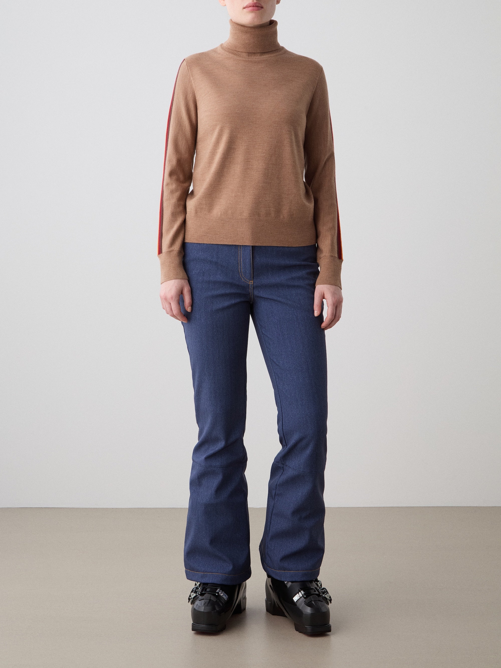 A person stands against a plain background wearing the We Norwegians FreeBird TurtleNeck women’s sweater with red-striped sleeves, blue jeans, and black platform shoes. Their arms are relaxed at their sides.