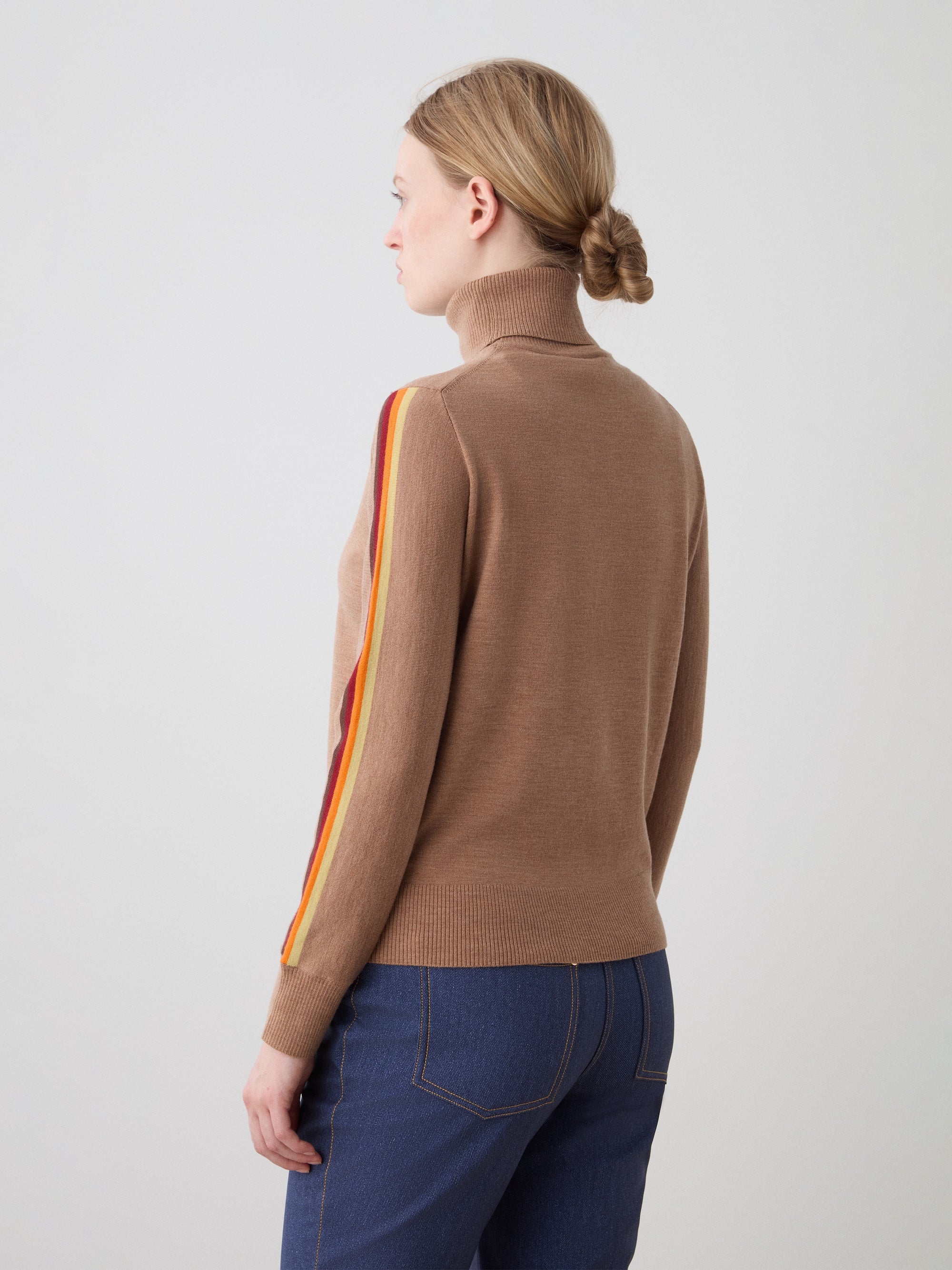 A woman with blonde hair in a low bun is wearing the We Norwegians FreeBird Turtleneck women sweater, featuring colored stripes on the sleeve, paired with blue jeans. She faces away, showing her left side against a plain gray background.