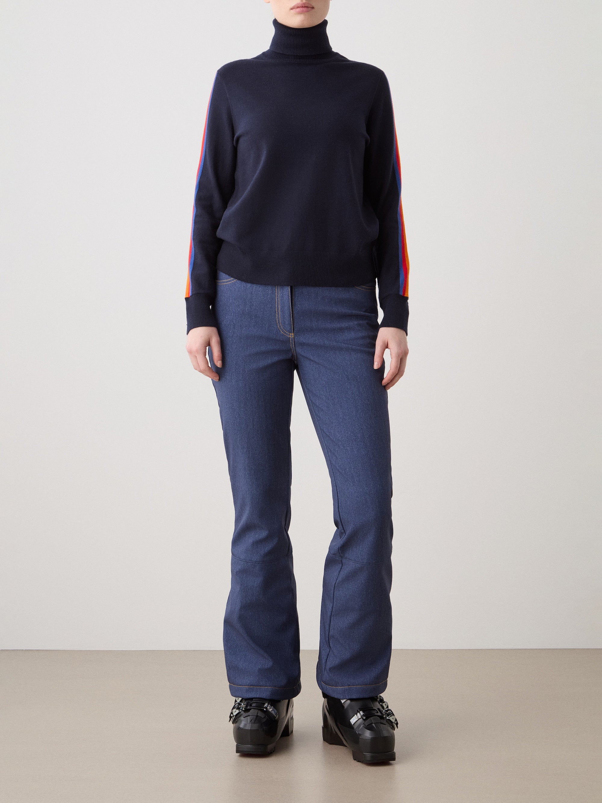 A person stands against a plain background wearing the We Norwegians FreeBird Turtleneck sweater with colorful striped sleeves, blue jeans, and black platform shoes. Their face is not visible.