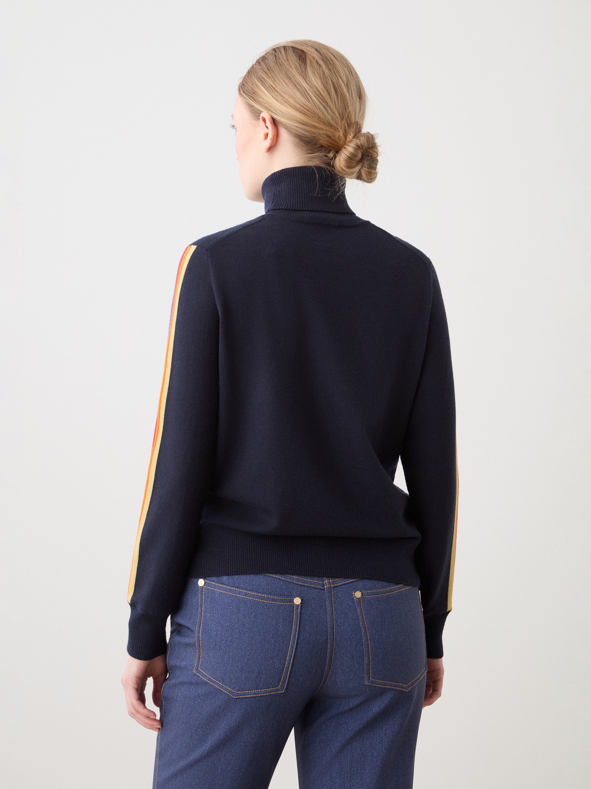 A person with blonde hair in a low bun is shown from the back wearing the We Norwegians FreeBird Turtleneck, a navy merino wool sweater with red, yellow, and cream striped sleeves, paired with blue jeans featuring gold stitching.