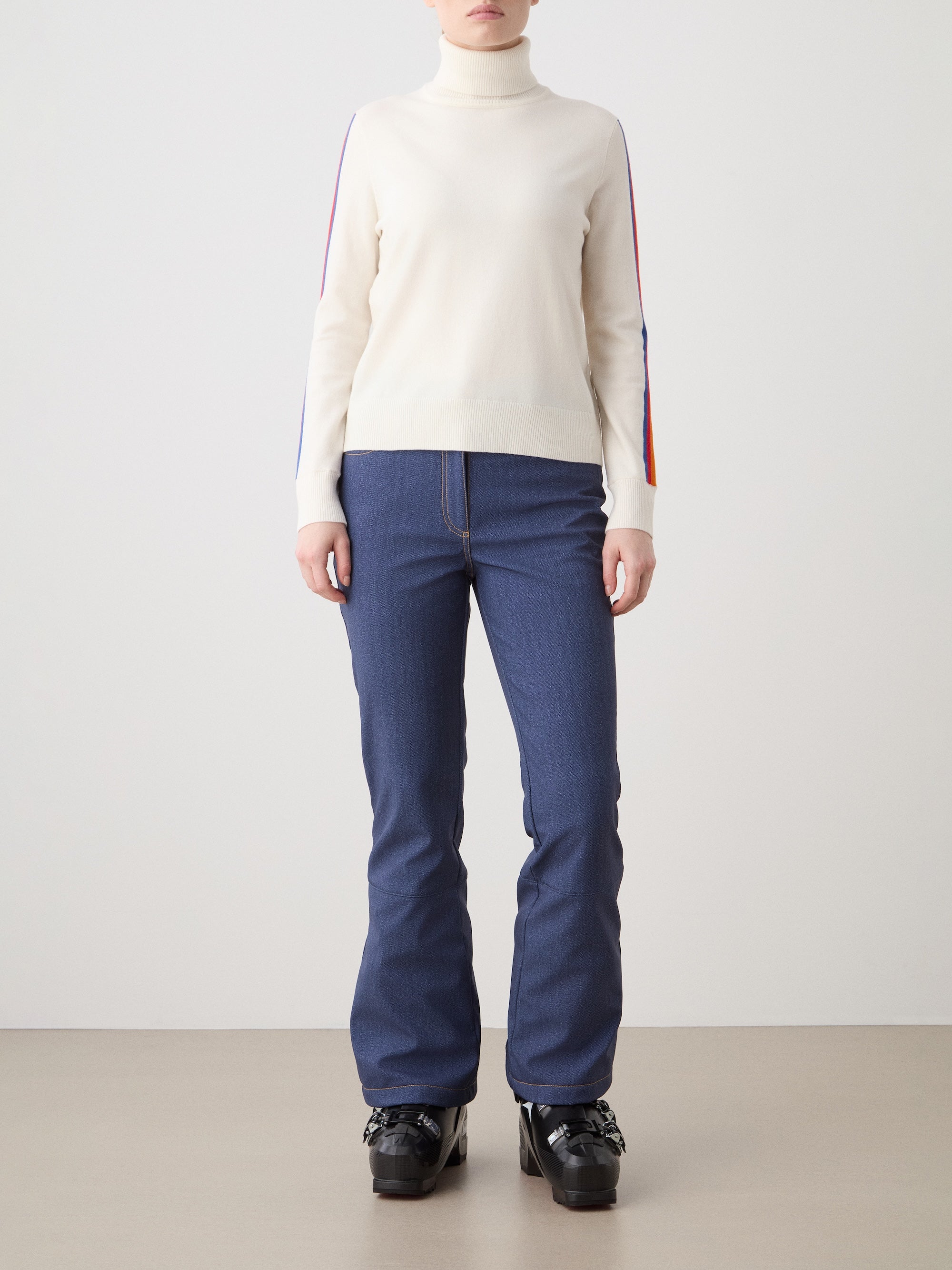 A person stands against a plain background wearing the We Norwegians FreeBird Turtleneck for women, featuring colored sleeve stripes, blue jeans, and black platform shoes. Their hands are by their sides; their face is not visible.