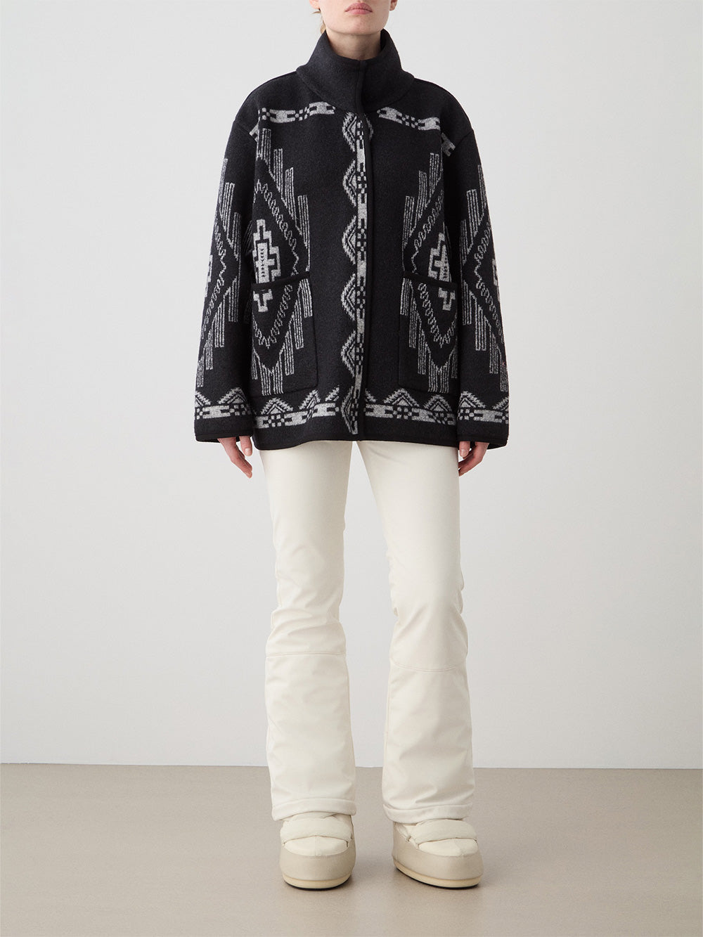 A woman stands against a plain background wearing the We Norwegians Kautokeino Jacket, featuring a black jacquard pattern with white geometric designs, a high collar, and loose fit. She pairs it with white pants and off-white boots, hands by her sides.
